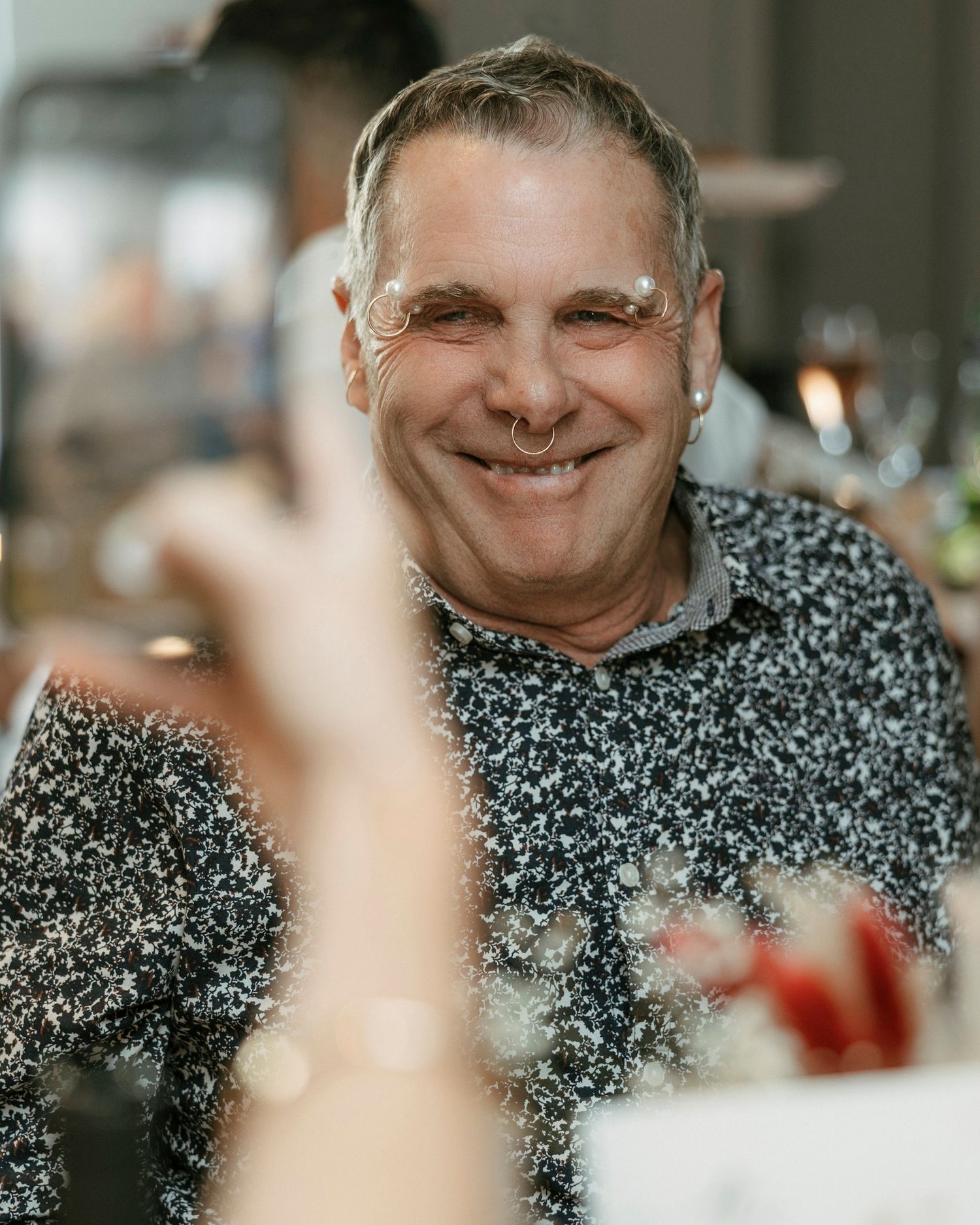 A joyful person wearing a speckled sweater sits at a table in a restaurant with a warm smile.