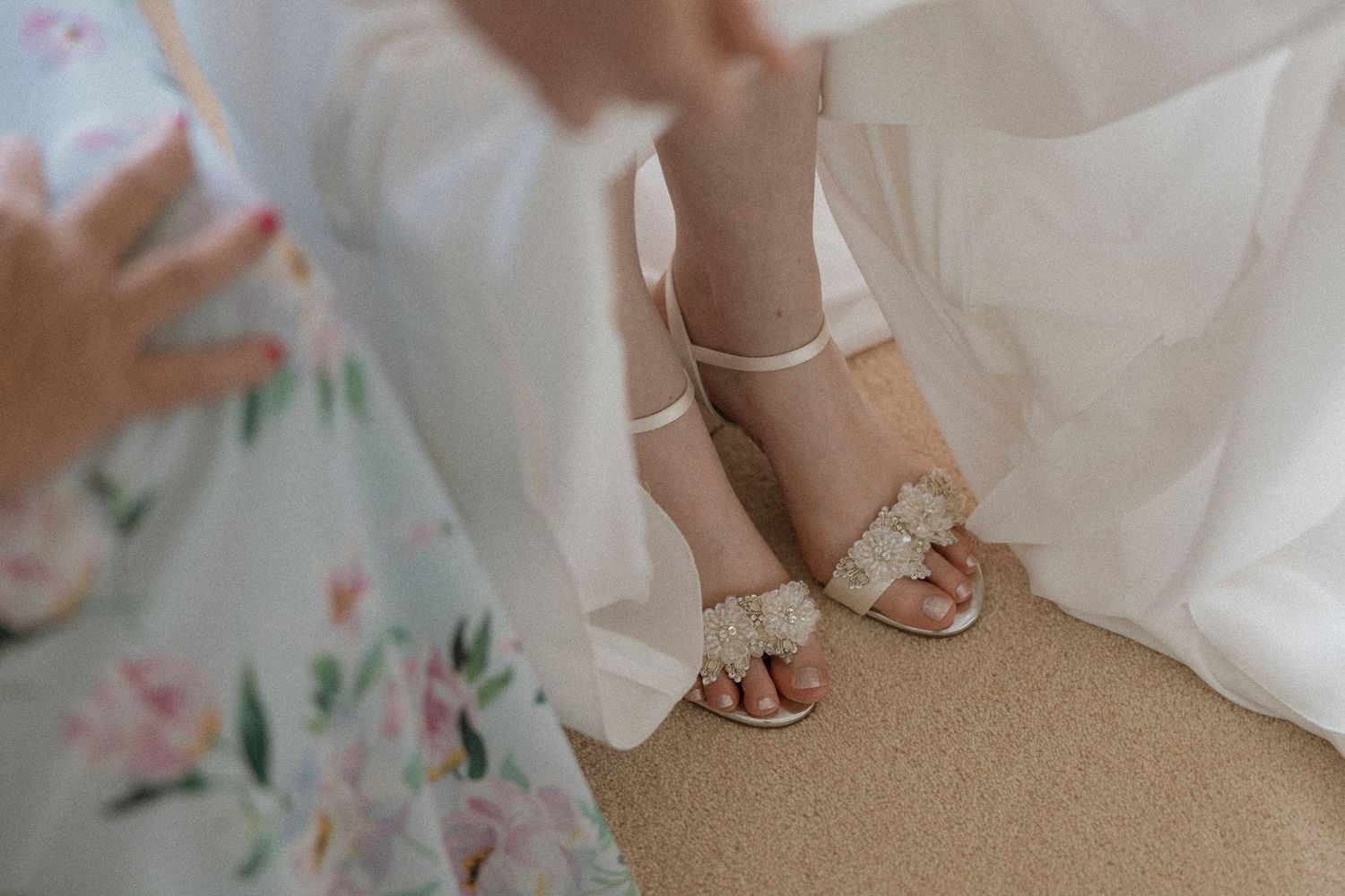 Close-up of a bride wearing elegant floral embellished wedding sandals beneath a flowing white bridal gown.