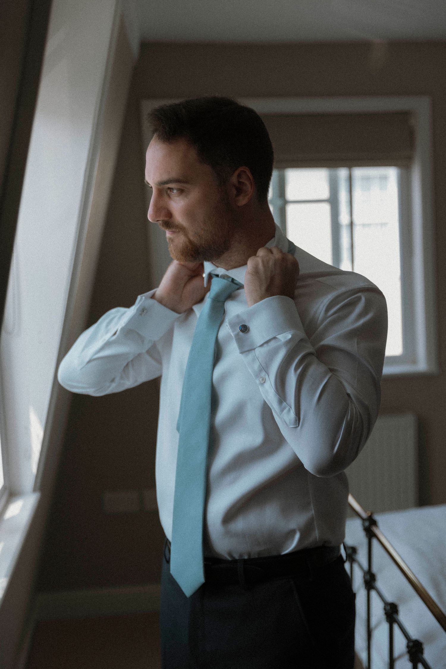 Man in white dress shirt adjusting light blue tie near window, getting ready for formal event.