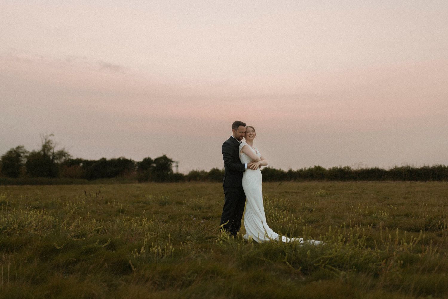 Bride and groom embracing in a green field at sunset, with a pink sky backdrop on their wedding day.