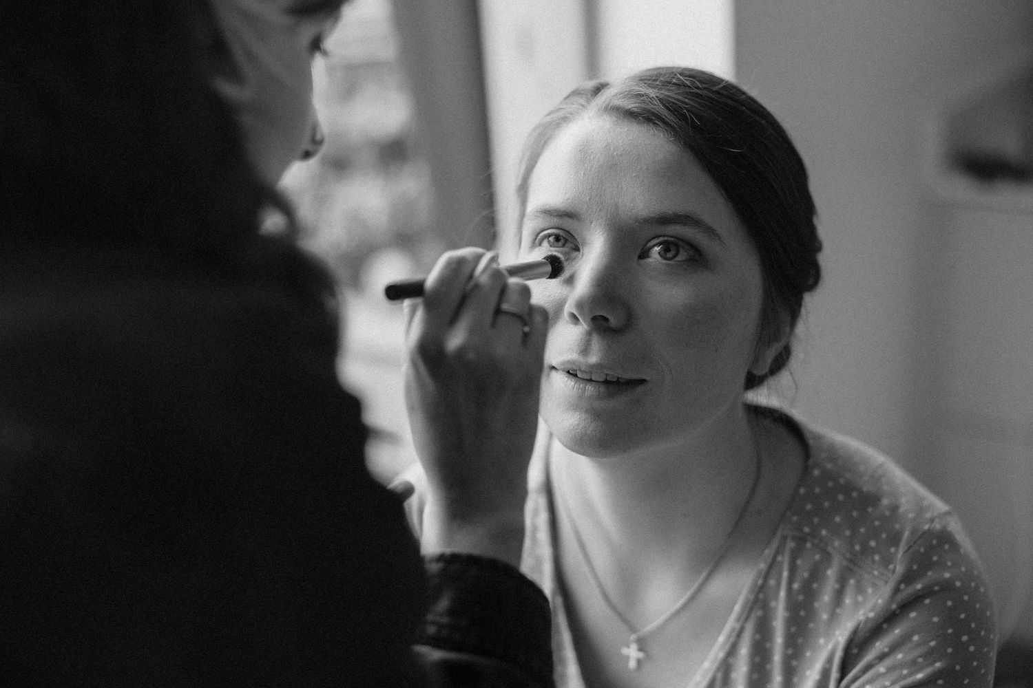 Black and white photo of a makeup artist applying eye makeup to a young woman wearing a necklace.