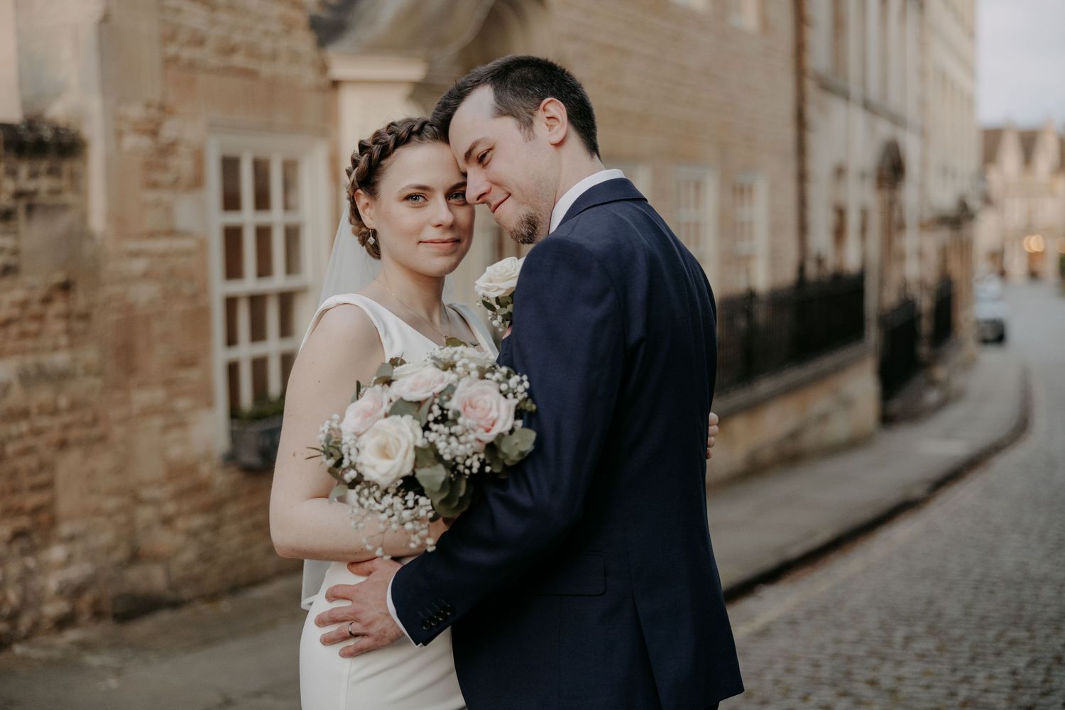 A bride and groom share a romantic embrace on a cobblestone street, she holds a pink and white bouquet.