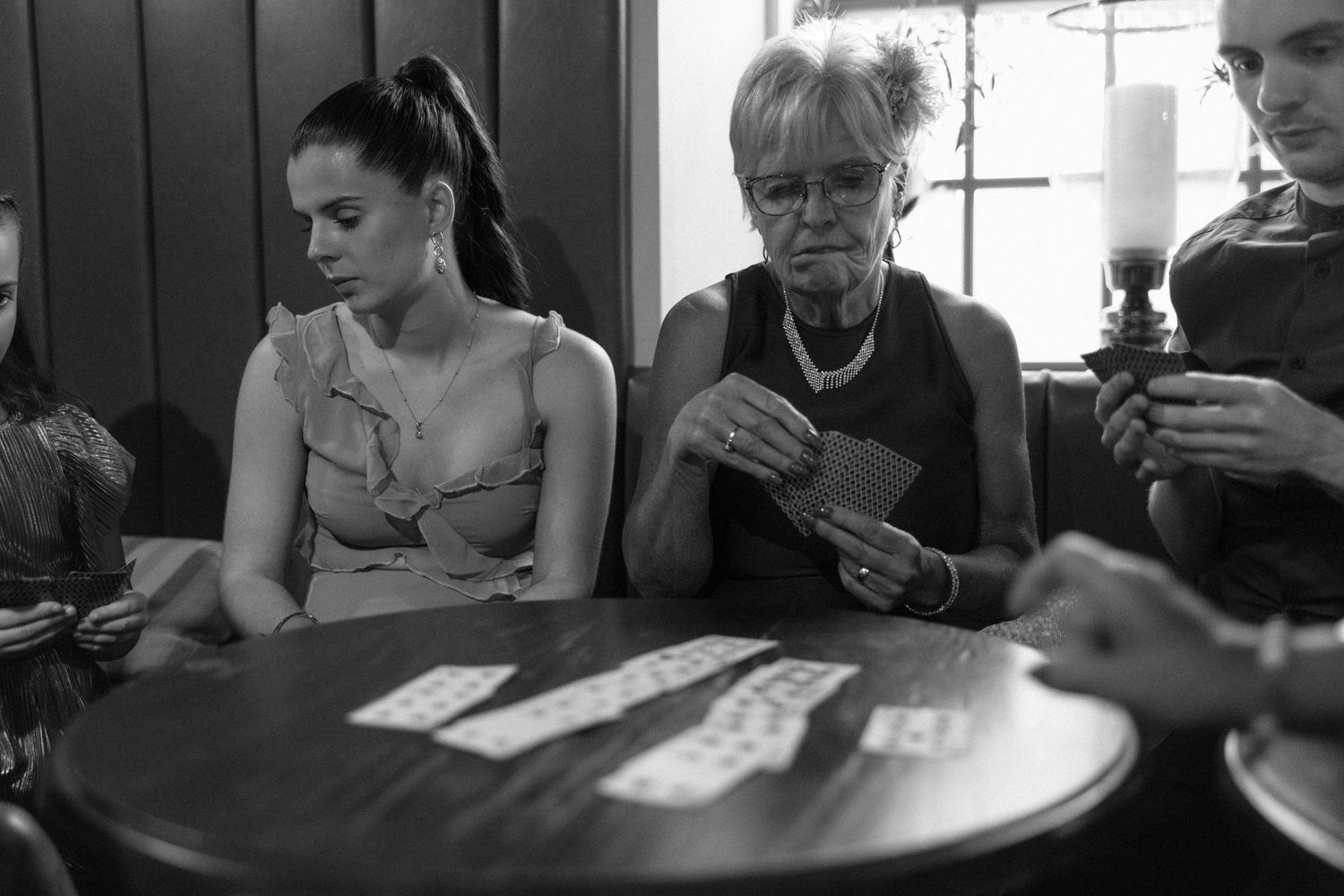 Black and white photo of people playing cards at a round table in a casual indoor setting.
