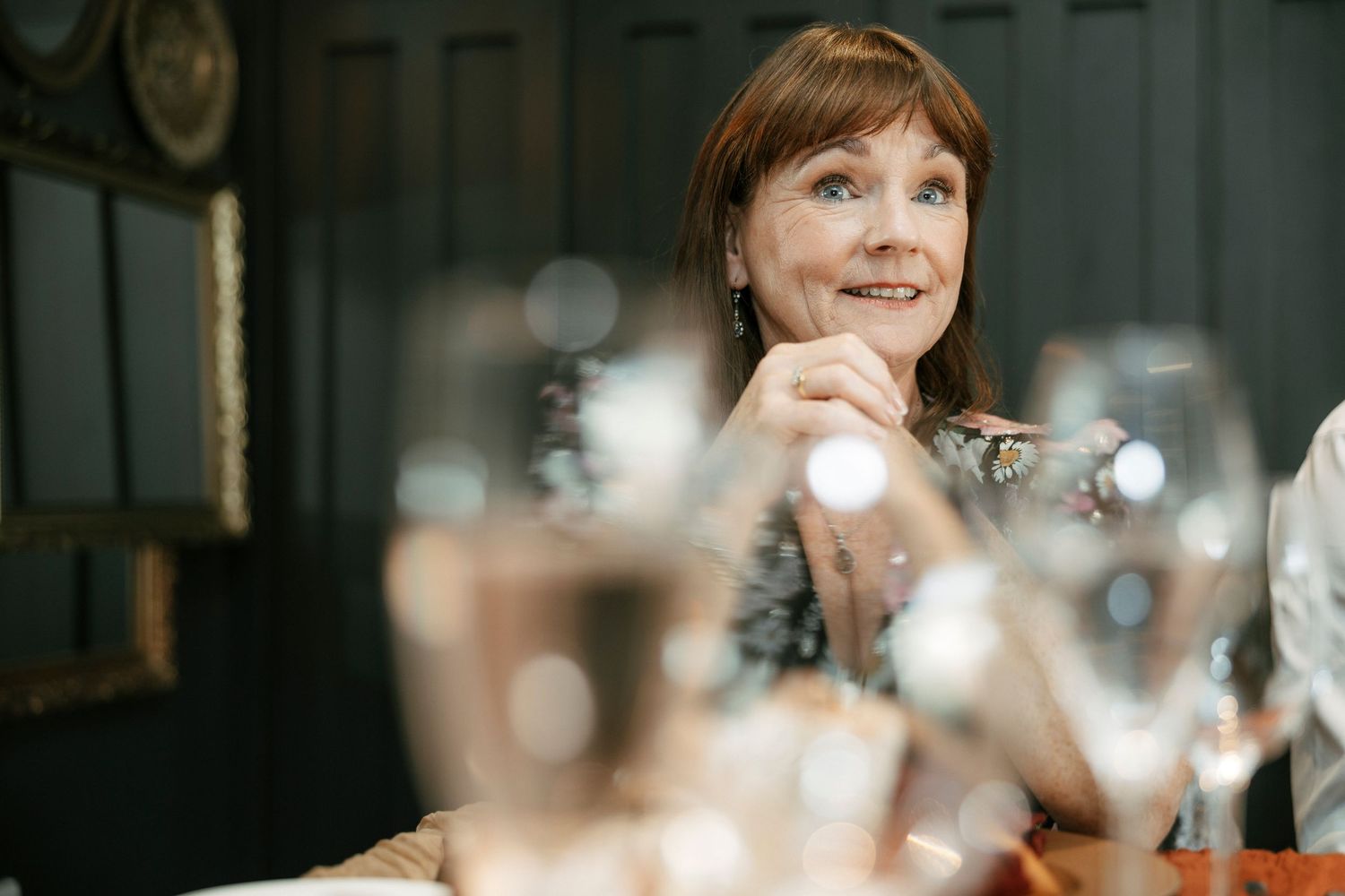A smiling woman with red hair sits at a dining table surrounded by elegant wine glasses in a dark interior.