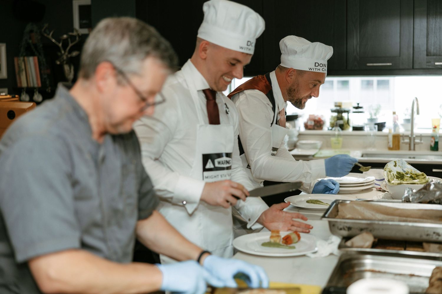 Three professional chefs in white uniforms and aprons plating gourmet dishes in a modern restaurant kitchen.