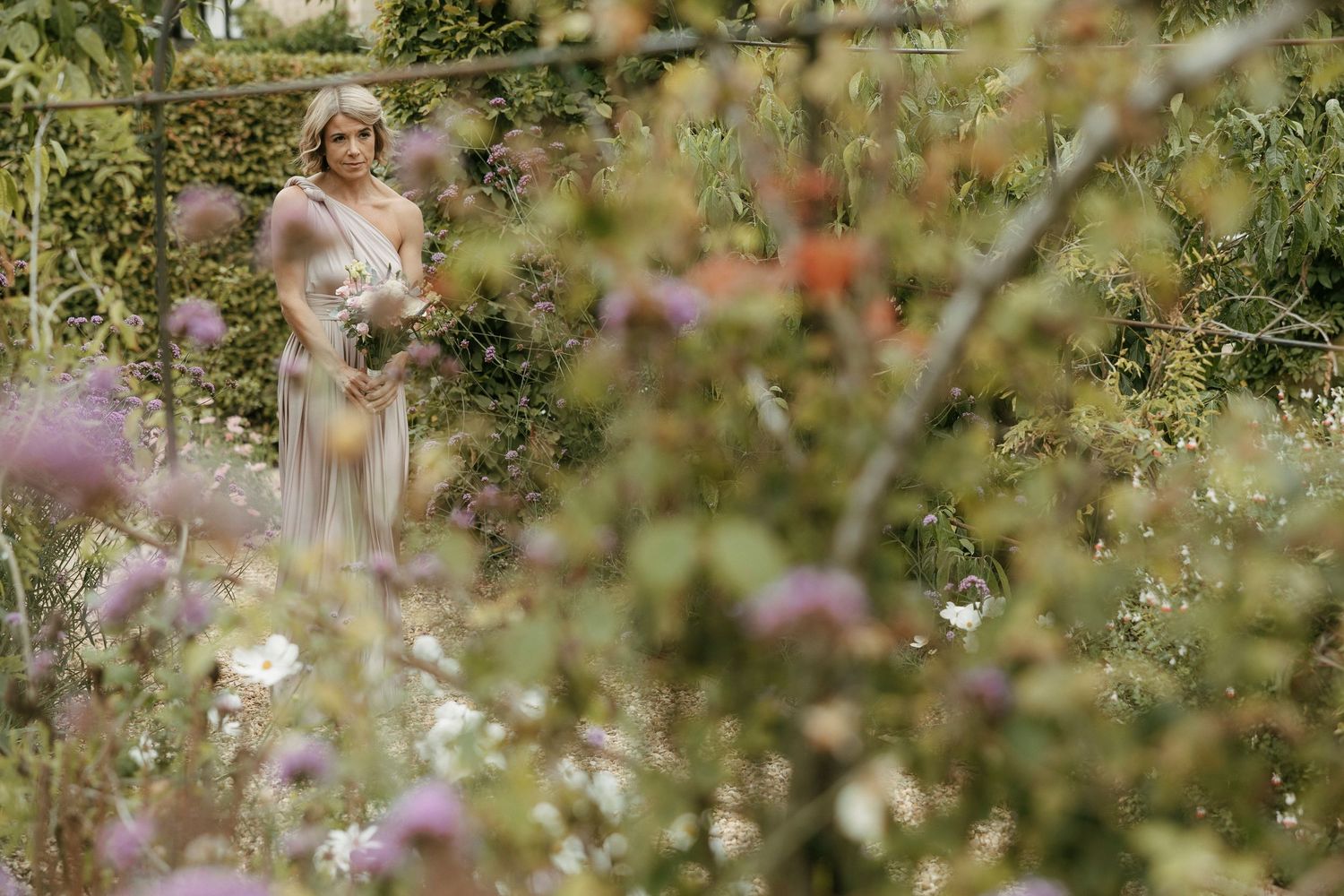 A woman in an elegant gown stands among lush garden blooms, surrounded by wildflowers in soft purple and white hues.