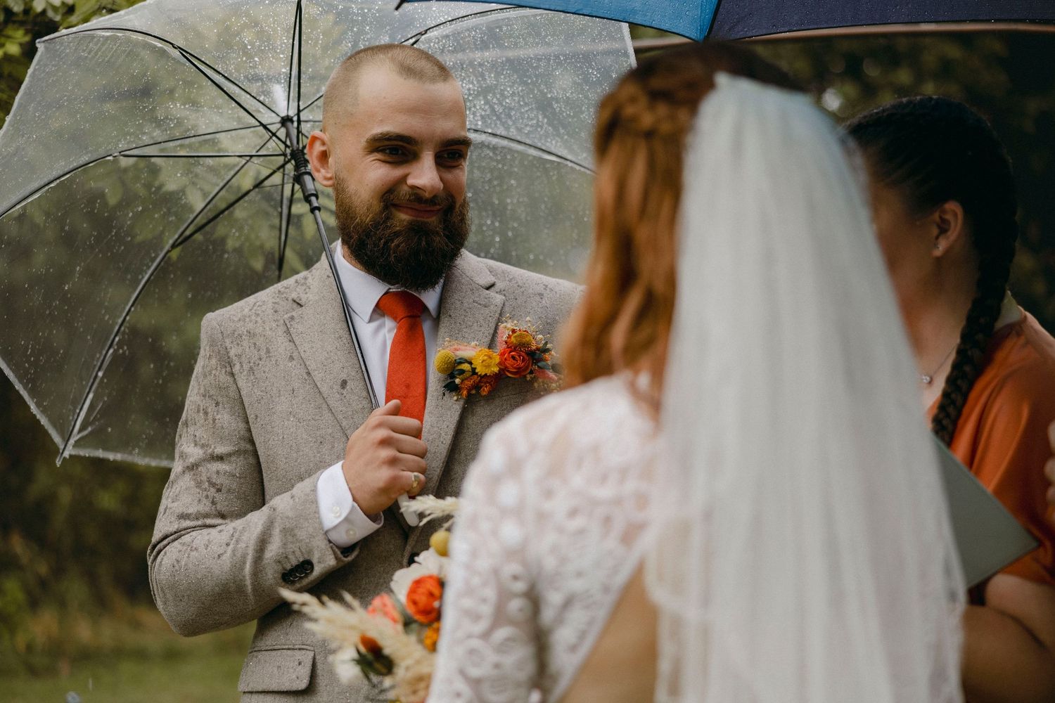 Groom in gray suit with orange tie holds clear umbrella, smiling at bride in white lace dress during outdoor rainy wedding ceremony.