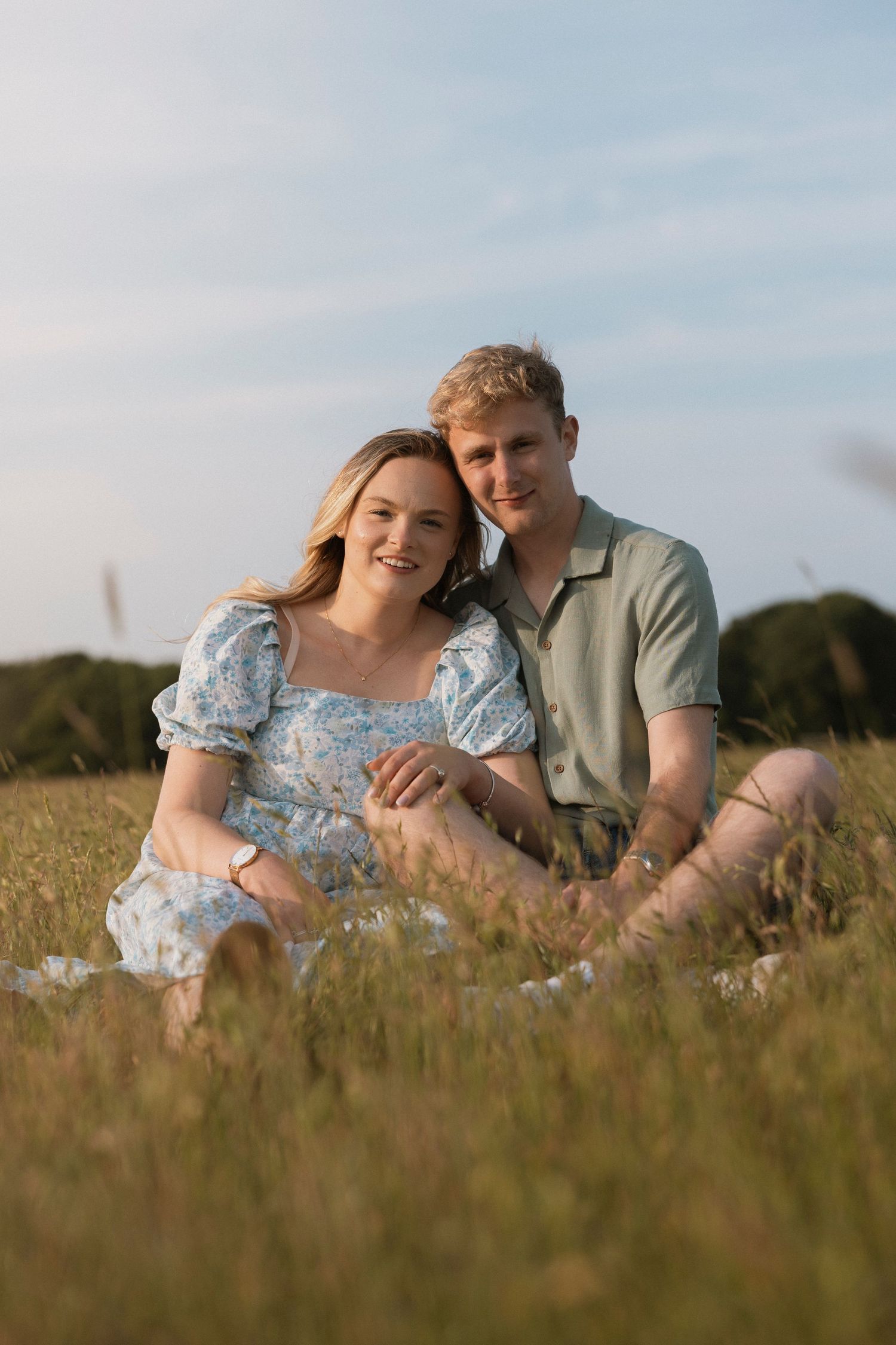 Couple sitting in a sunny grass field, woman in floral dress and man in sage shirt, smiling at camera.