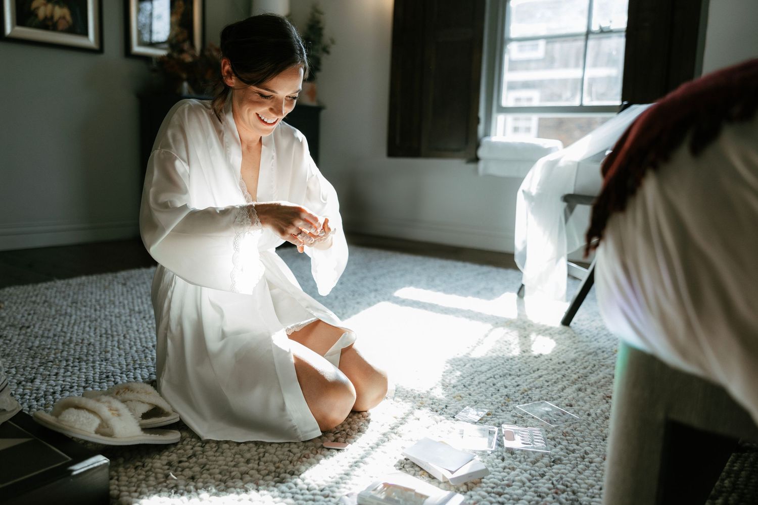Bride in white robe kneeling on floor putting on wedding garter in sunlit room.