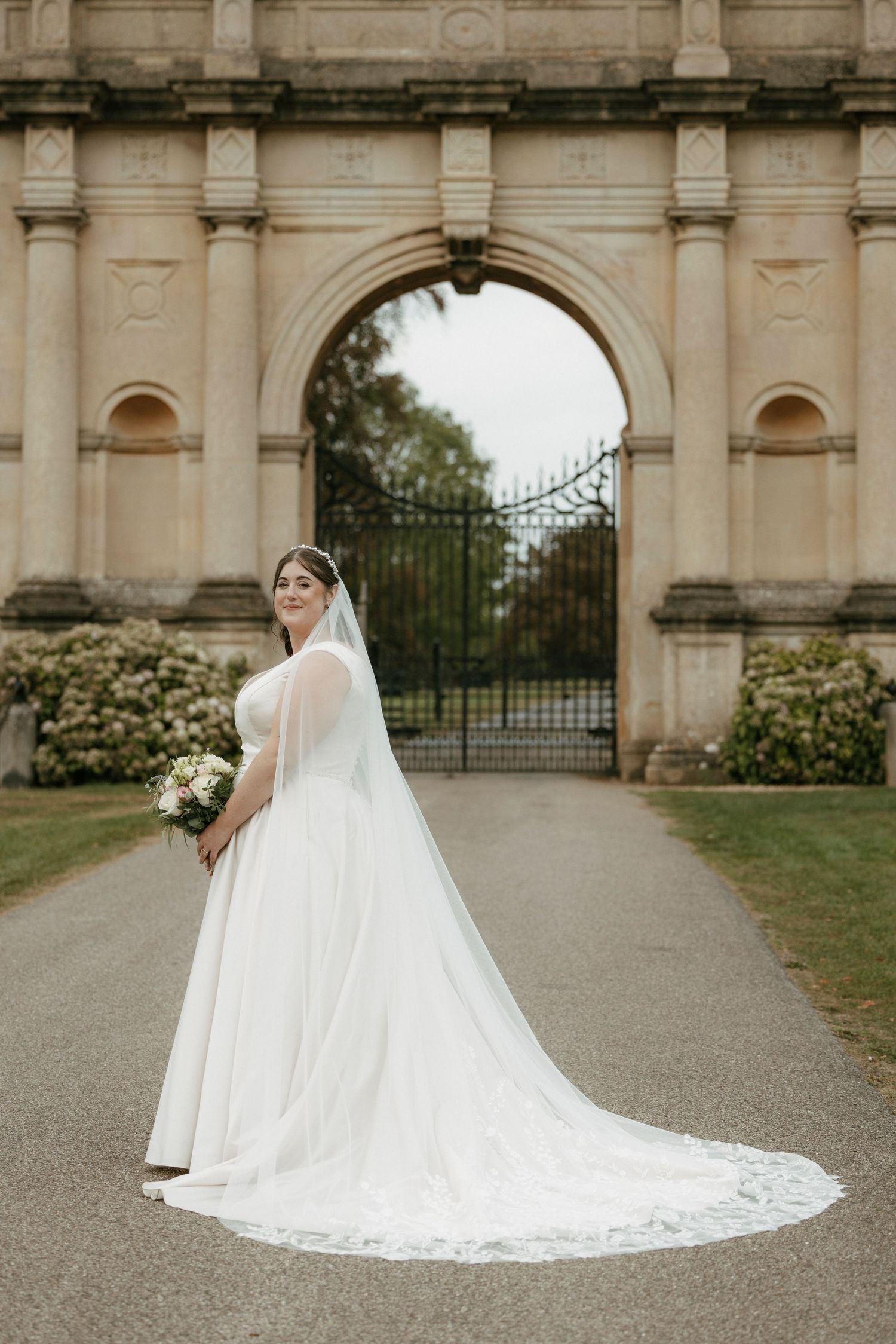 Bride in a long white gown with cathedral veil holds bouquet before ornate stone arch gate.