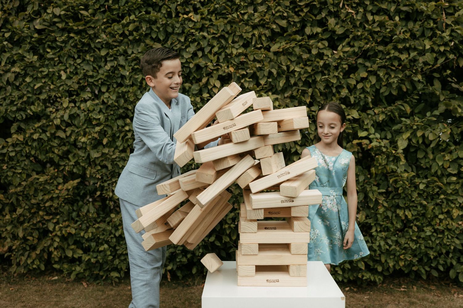 Two children playing giant Jenga outdoors, laughing as wooden blocks tumble from the tall tower.