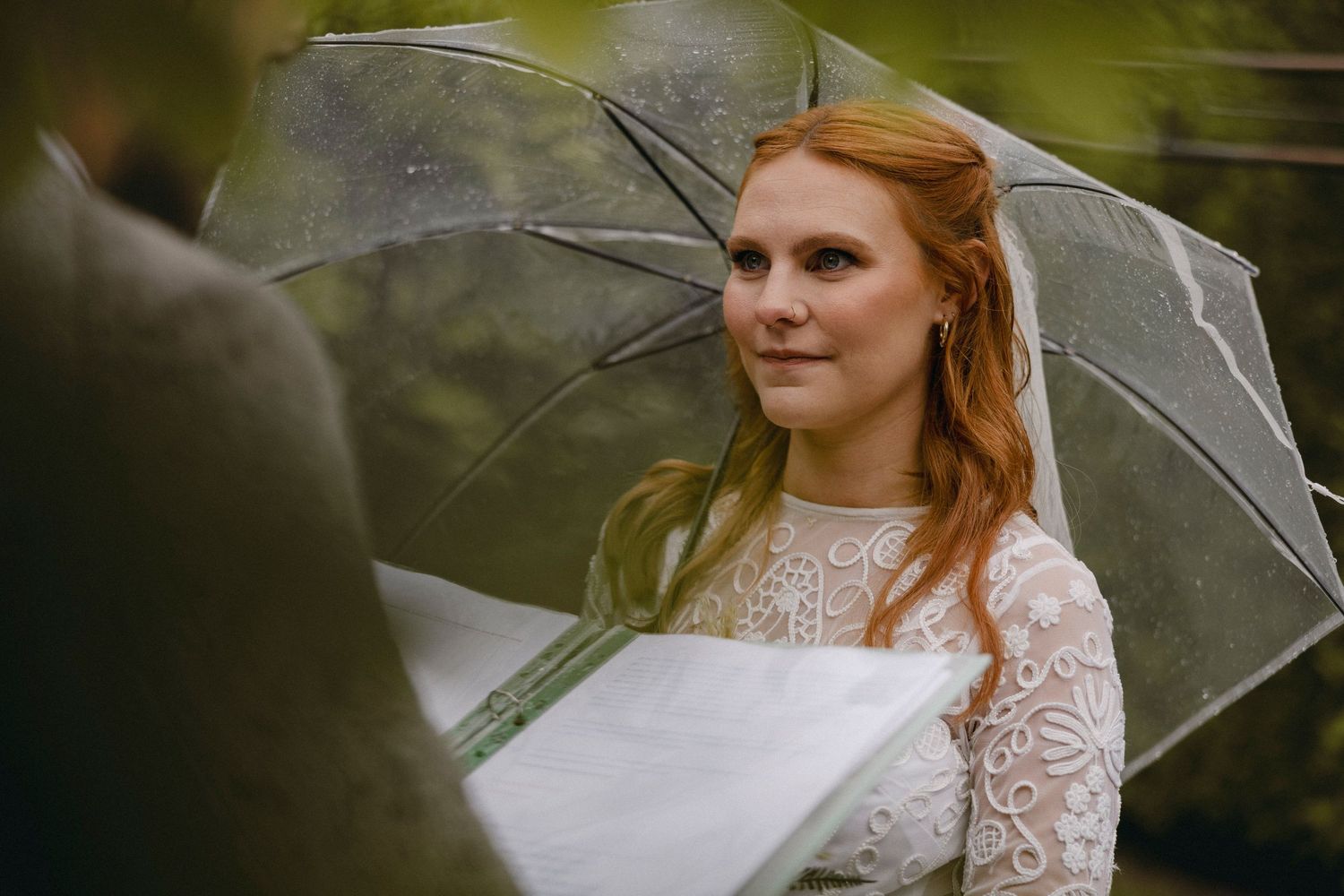 A red-haired bride in a lace wedding dress holds a clear umbrella outdoors on a rainy wedding day.