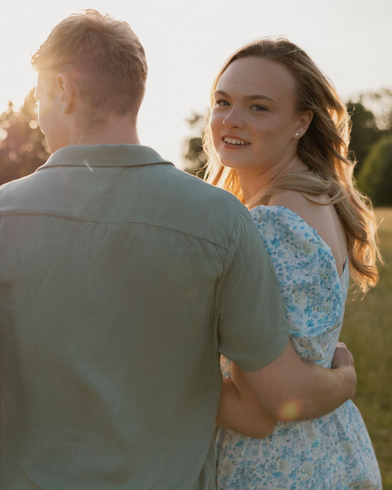 Romantic sunset photoshoot with couple walking in sunlight wearing blue floral dress and grey shirt in outdoor field.