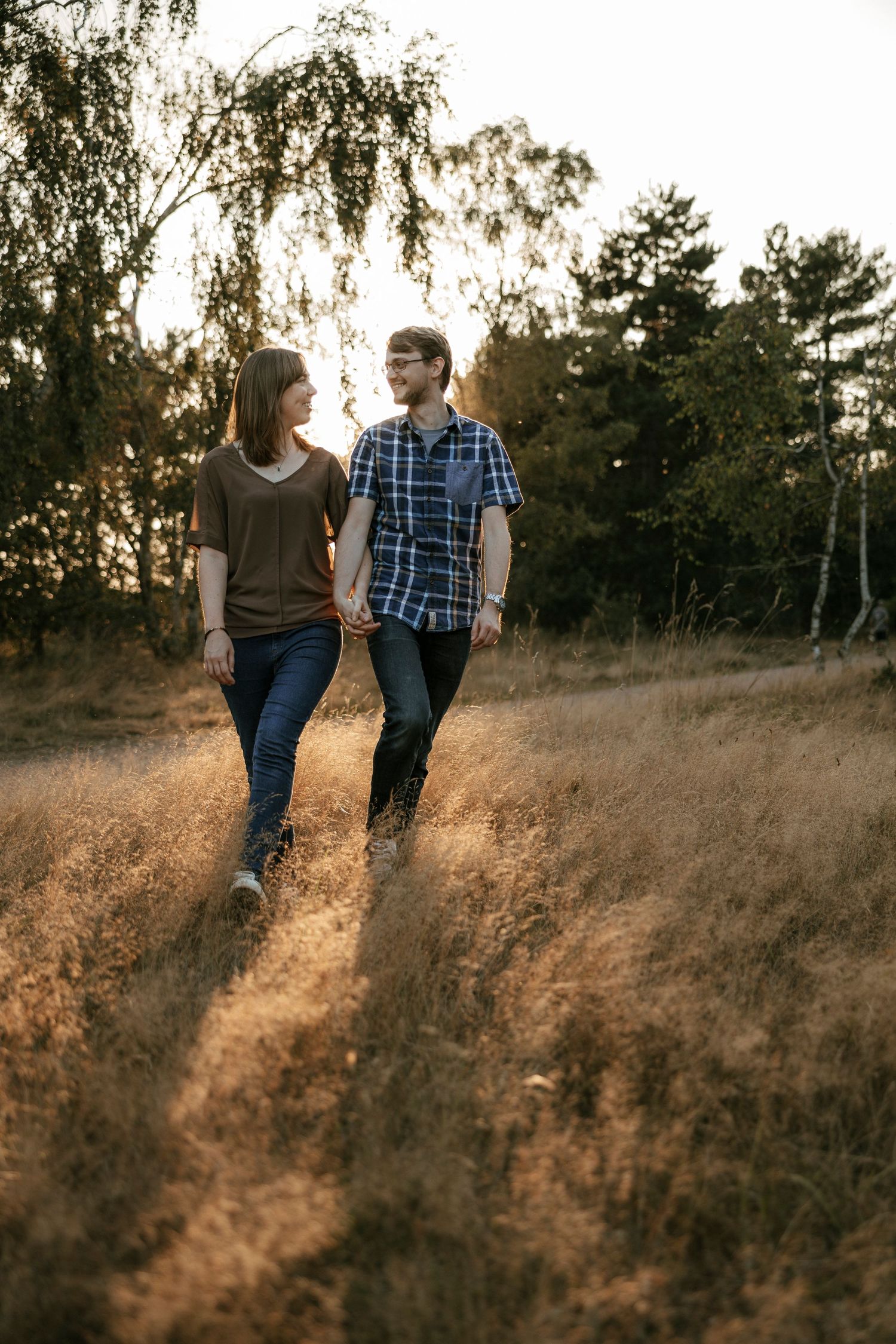 Couple holding hands walking through golden grass field at sunset with trees in background.