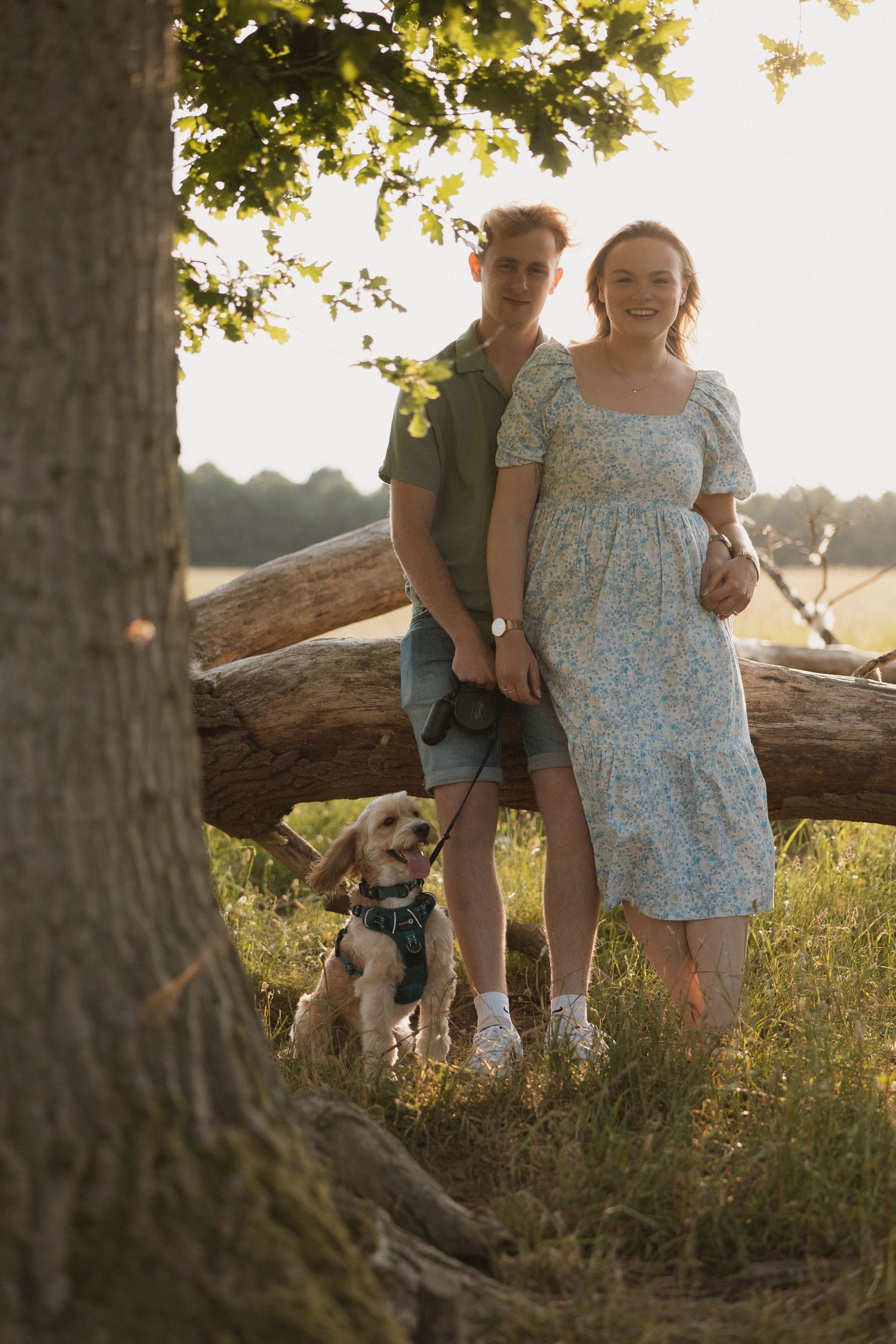 Couple with a golden Cockapoo dog leaning against a fallen log in a sunny meadow.