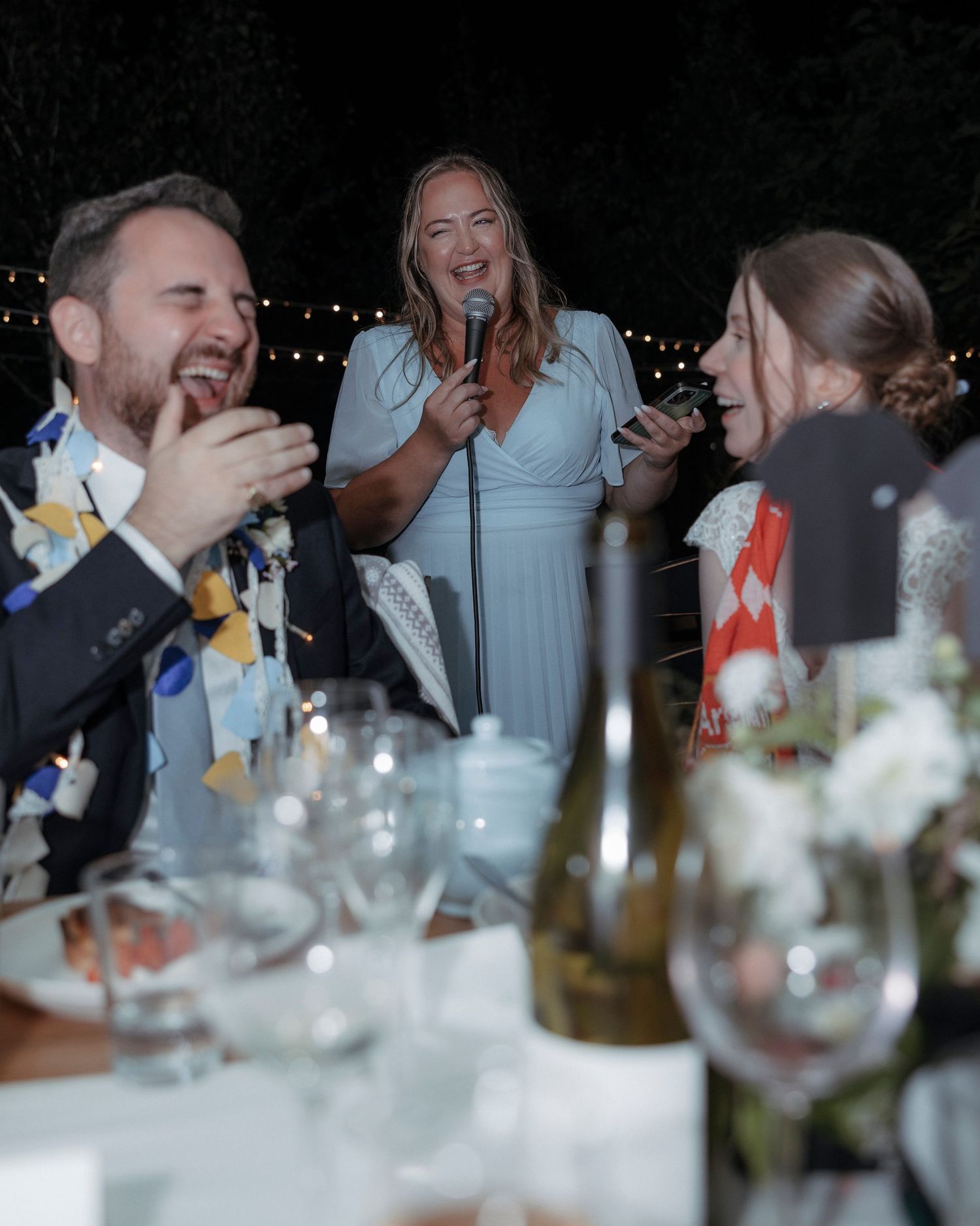 Guests laughing and singing with microphones at an outdoor wedding reception dinner at night.