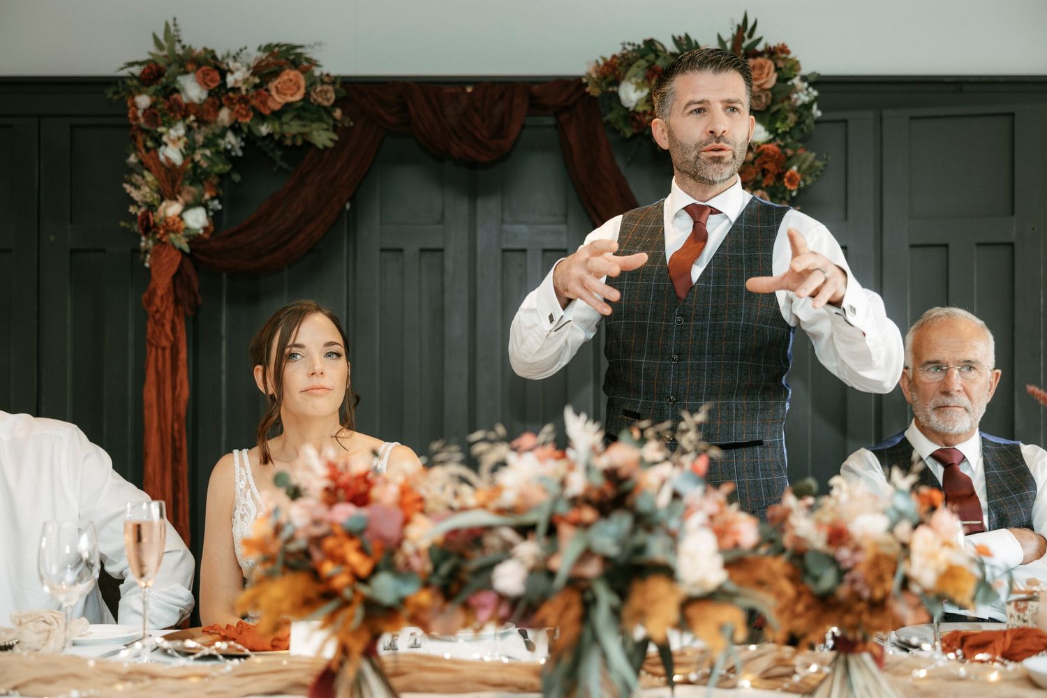A groom gives an animated speech at a wedding reception decorated with floral arrangements.