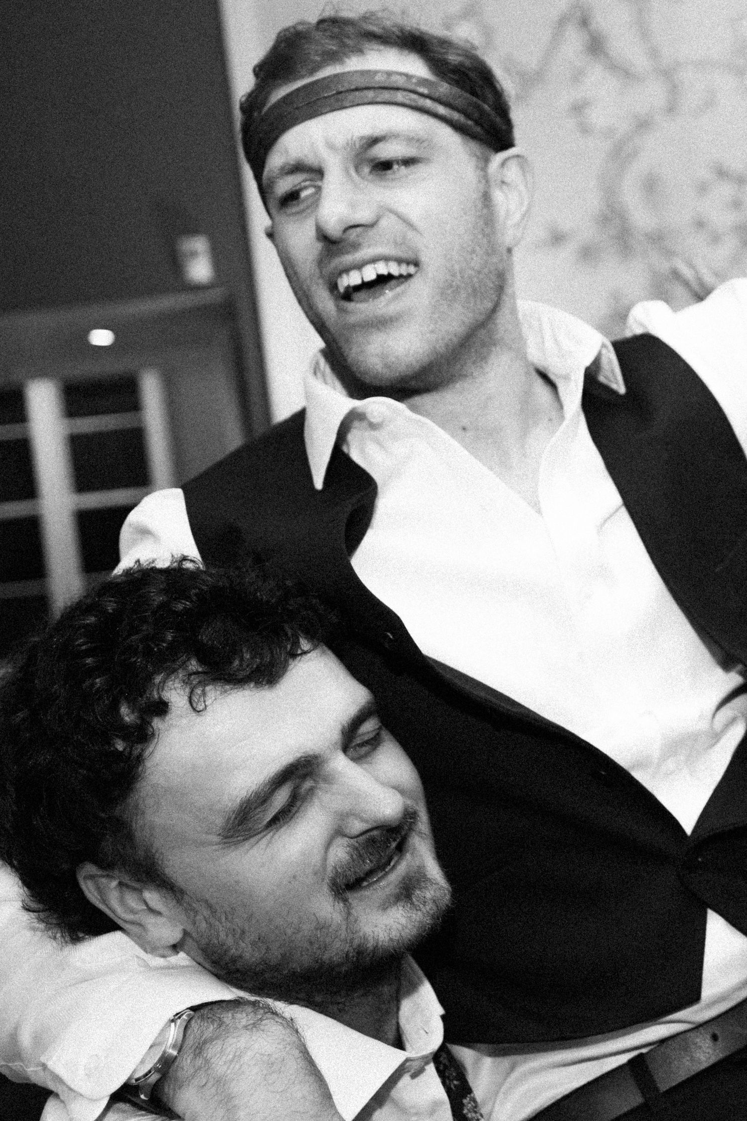 Black and white photo of two laughing men, one wearing a headband, sharing a joyful moment at a social event.