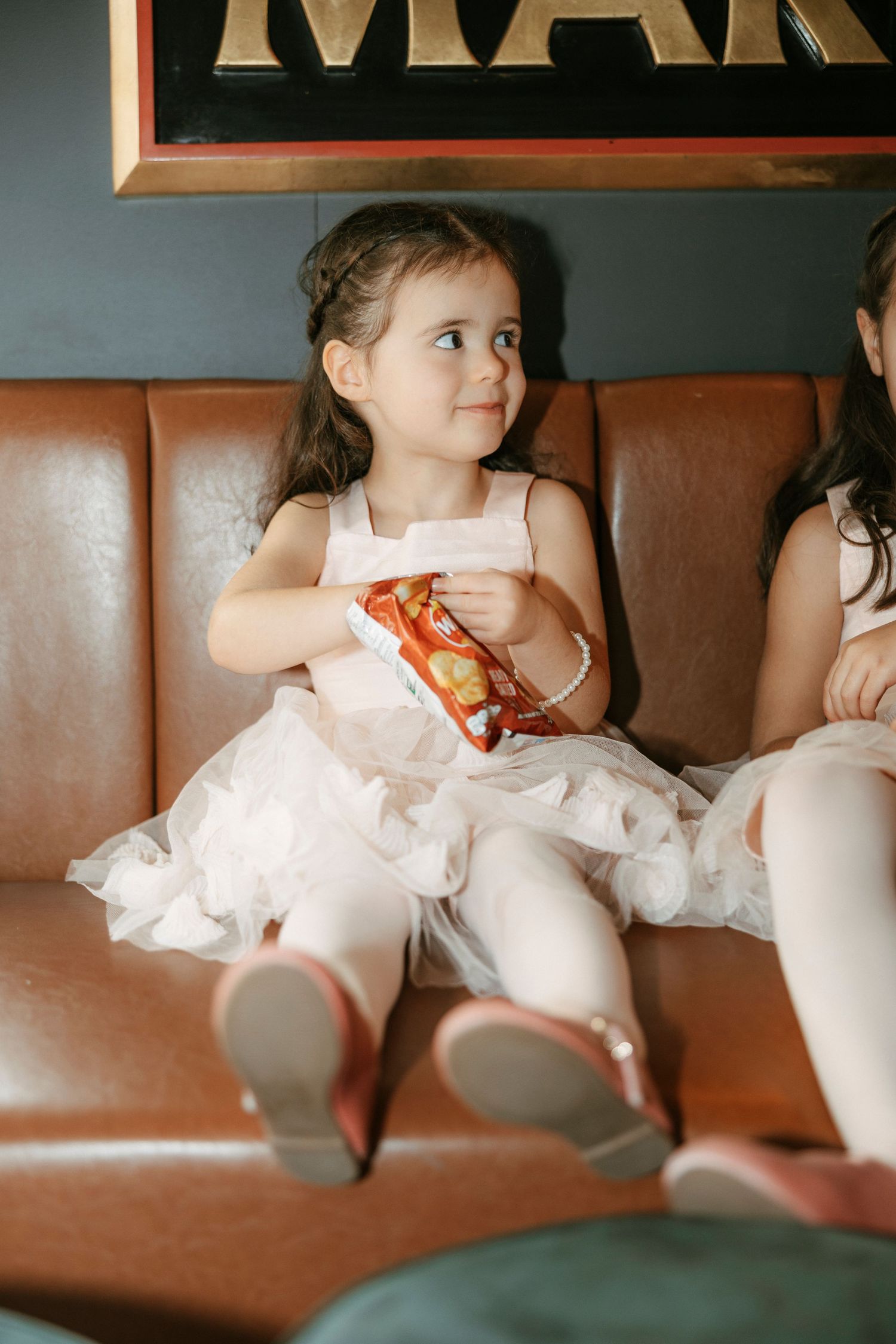 A child in a white dress sits on a brown leather booth holding a snack while looking to the side.
