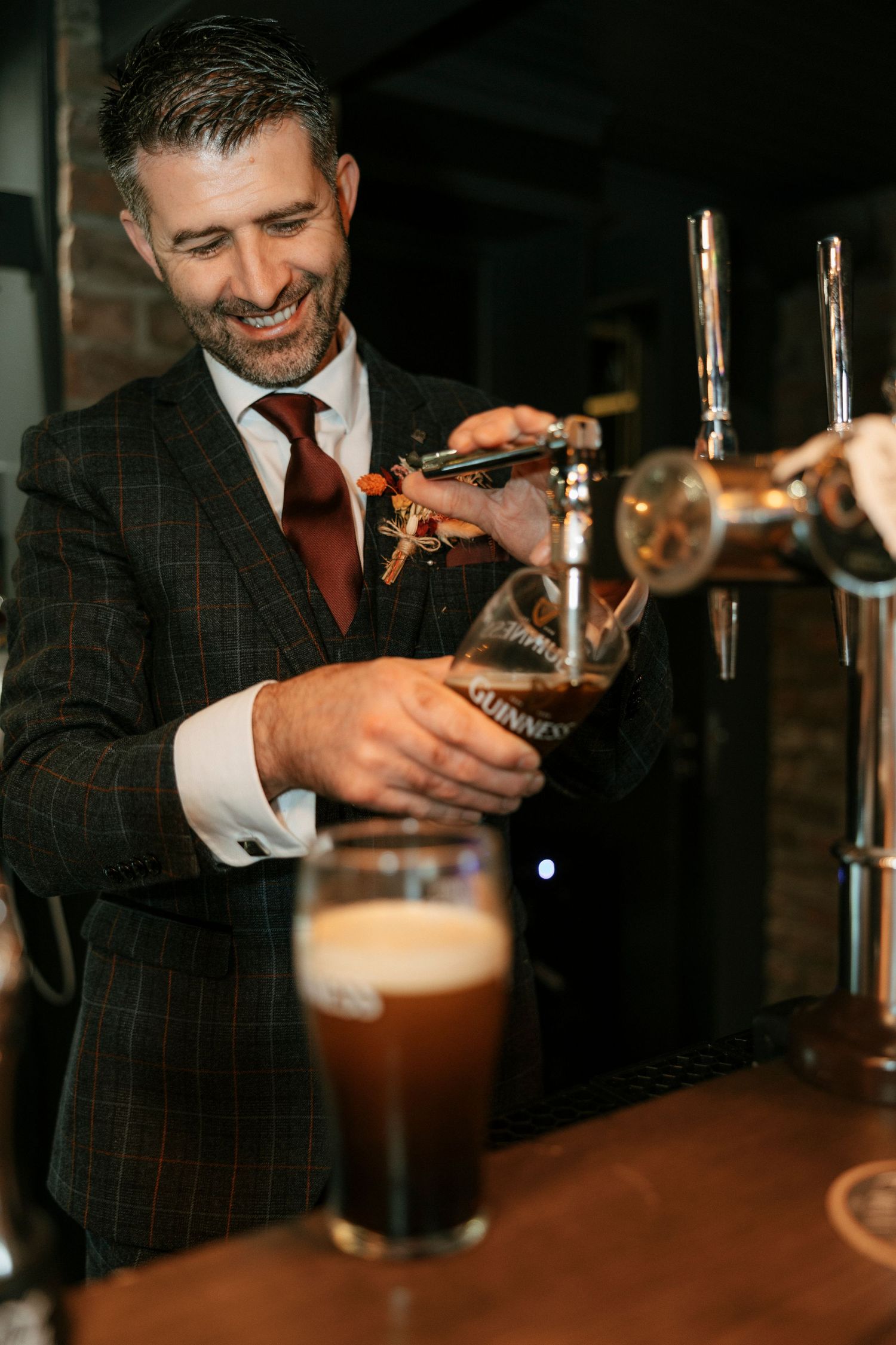 Groom in plaid suit pouring draft beer at wedding reception bar.
