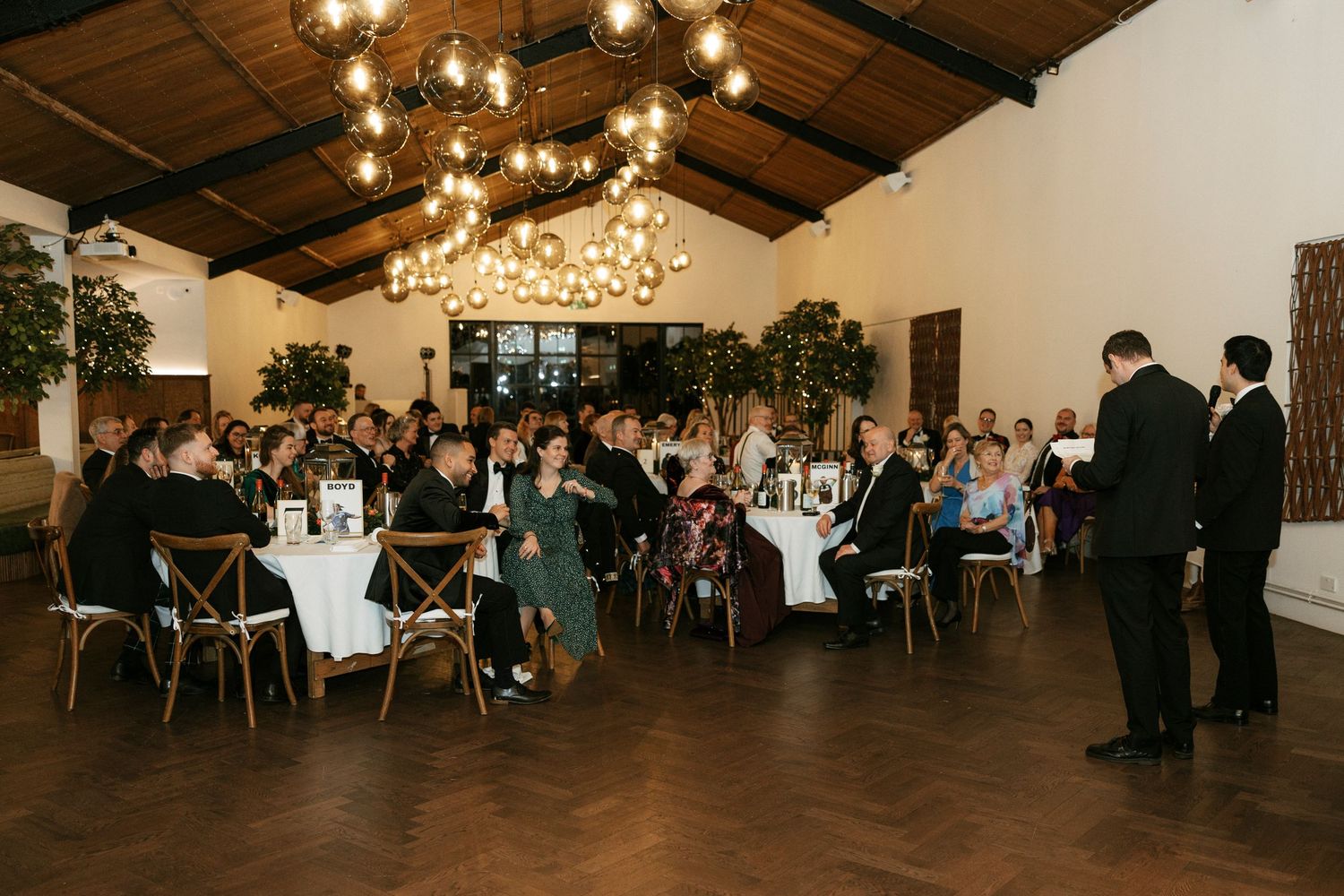 Elegant wedding reception in a rustic venue with pendant lights, round tables, and guests listening to a speech.