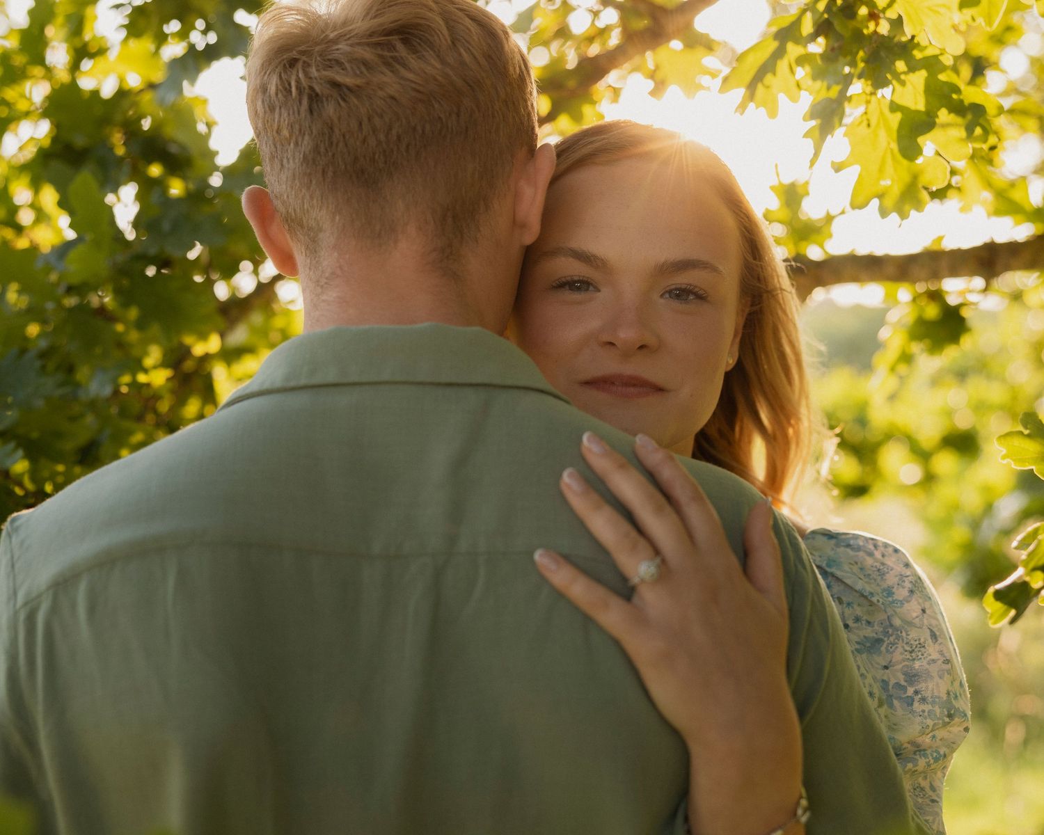 Couple embracing outdoors in golden sunlight near green grapevines, woman wearing engagement ring smiling warmly.