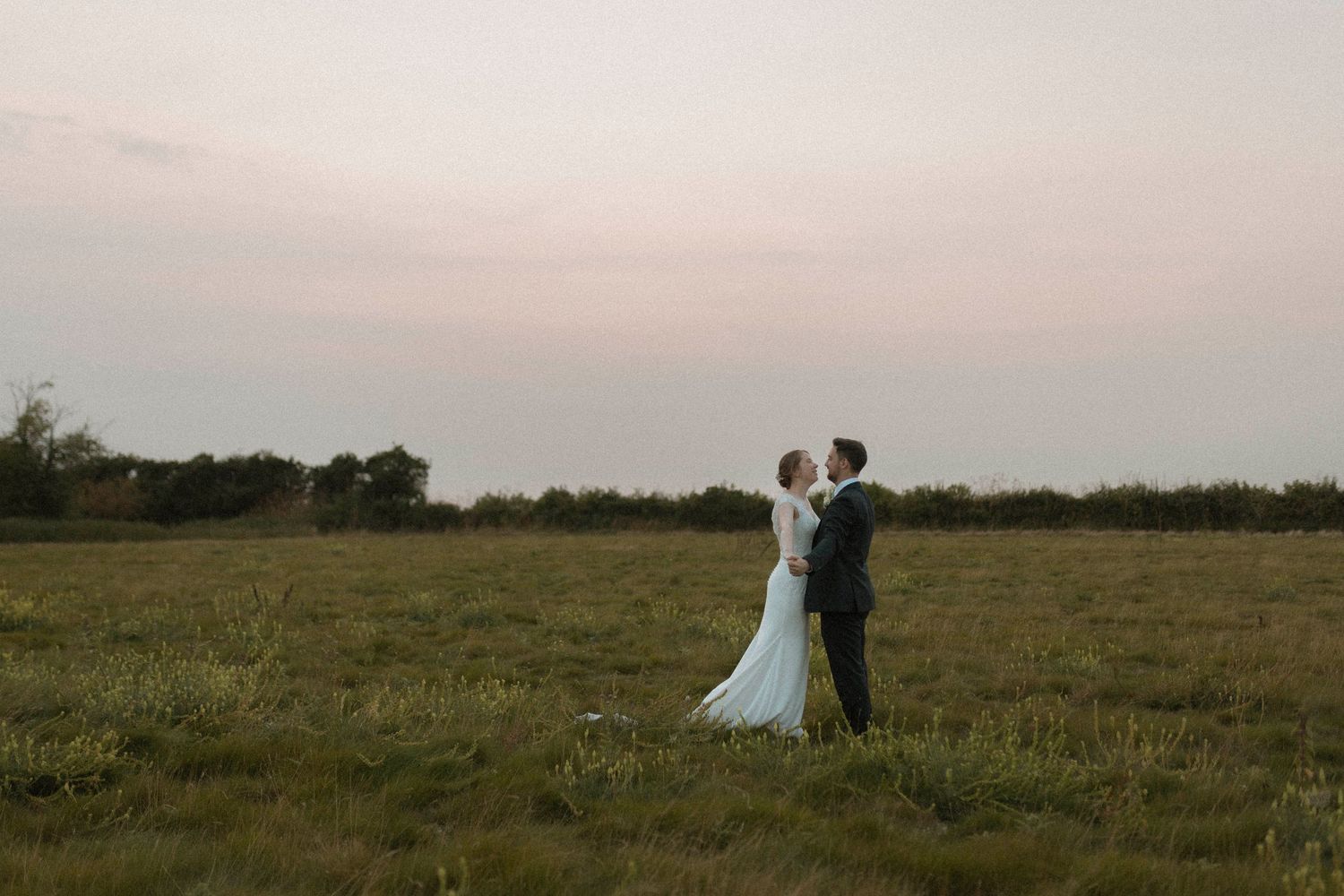 A bride and groom share a romantic embrace in an open green field at dusk during their outdoor wedding ceremony.