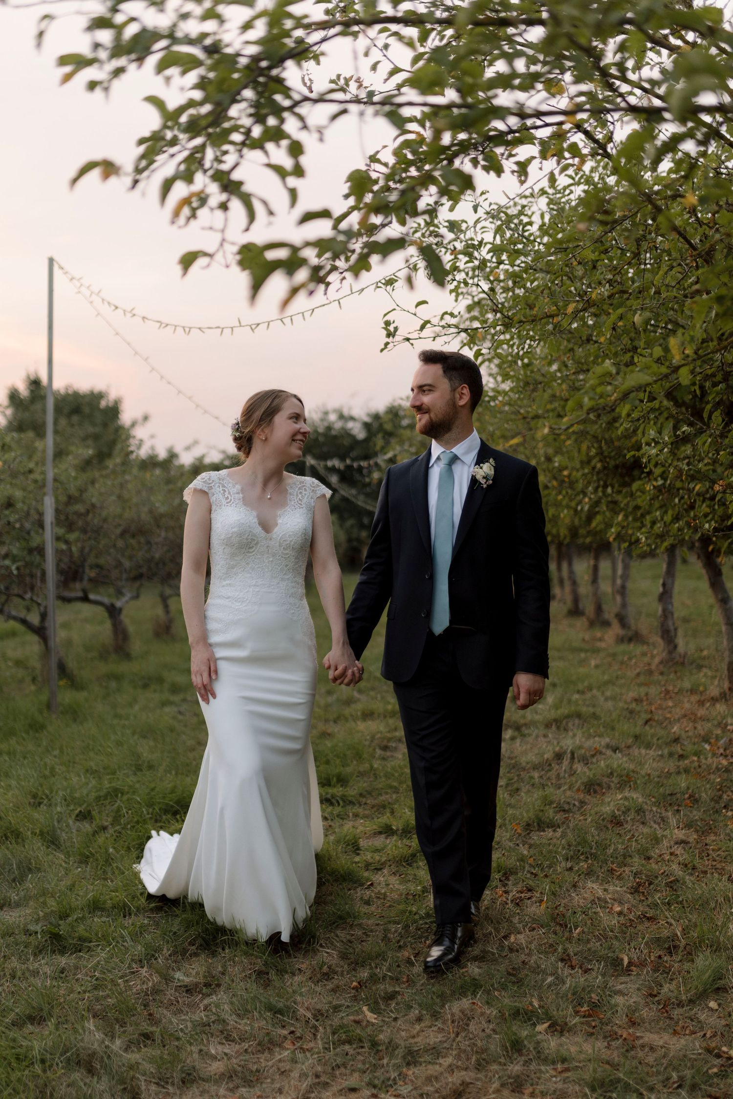 A bride and groom walk hand in hand through a lush orchard at sunset, smiling at each other on their wedding day.