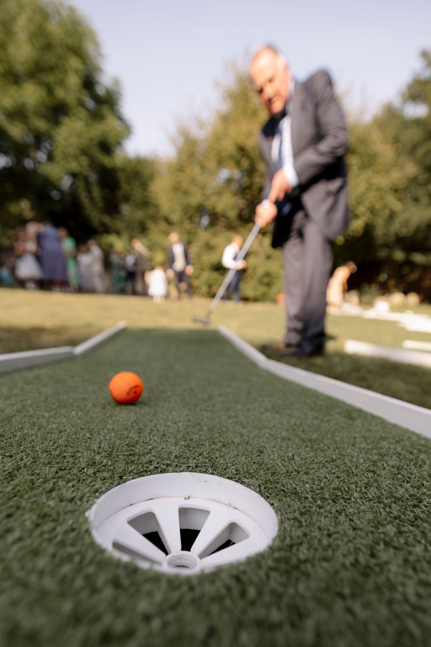 Man in suit playing mini golf, aiming orange ball toward white hole cup on artificial green turf outdoors.
