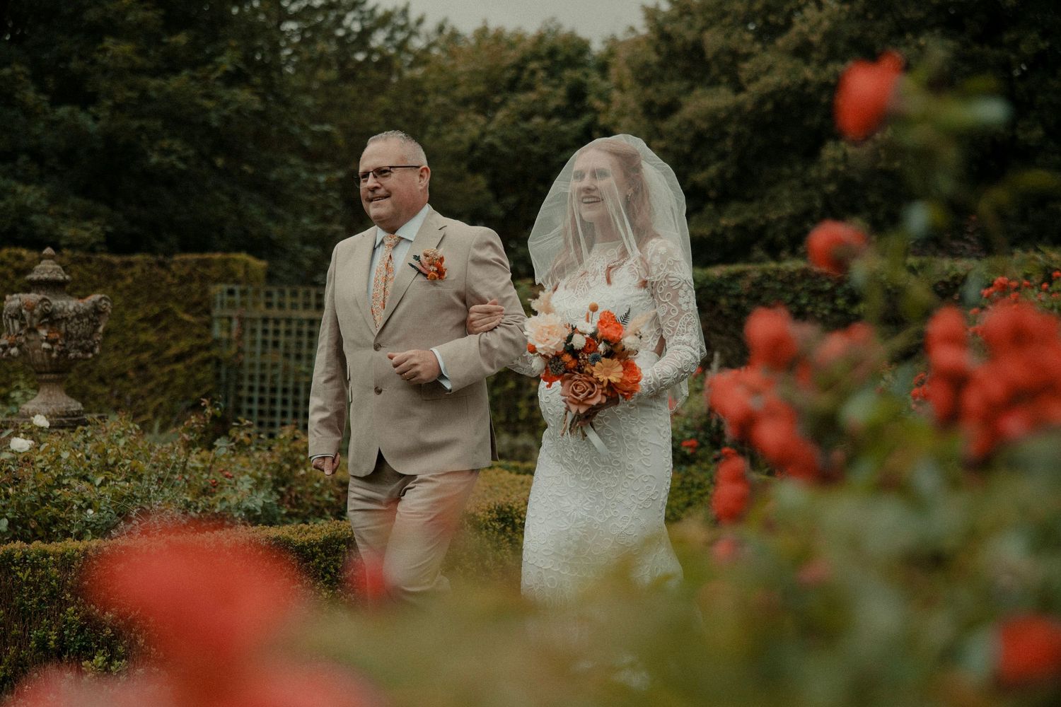 Bride and father walk through a rose garden, bride holding a vibrant bouquet in a lace gown with veil.