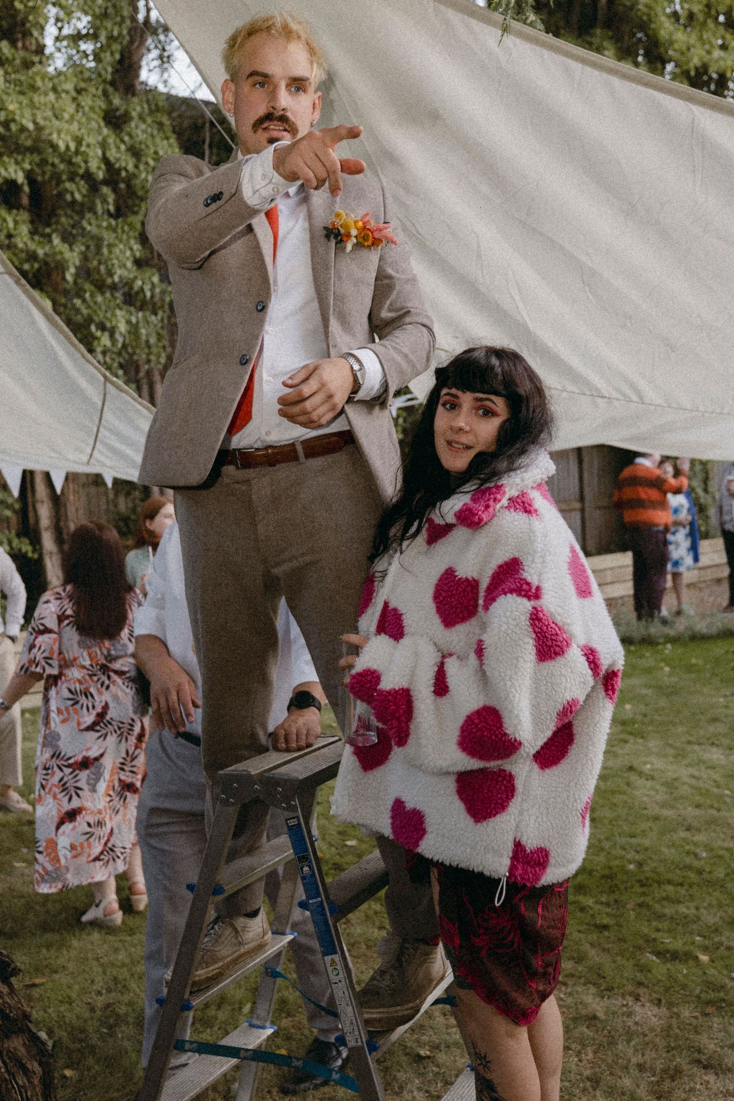 A man in a grey suit on a ladder points down at a woman in a white heart-patterned sweater at an outdoor wedding.