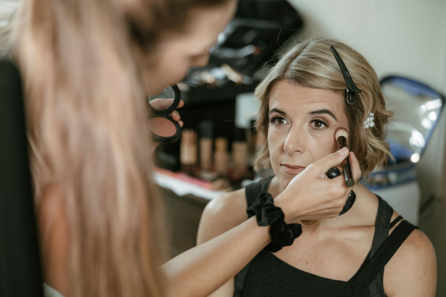 A makeup artist applies foundation to a blonde woman's face in a professional beauty salon setting.