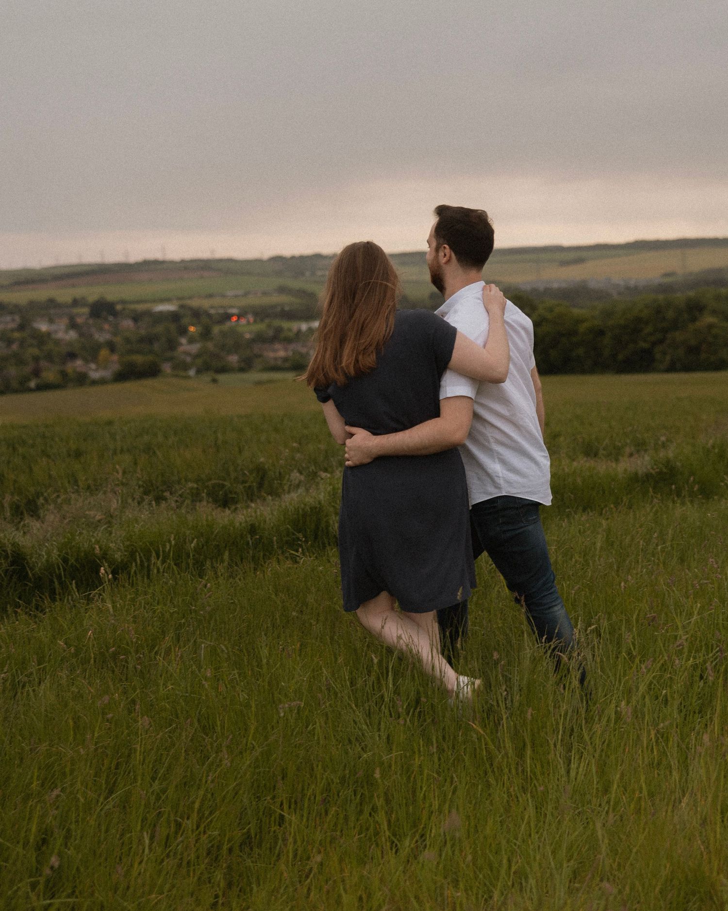 Couple embracing while walking through tall grass on a hillside at dusk.