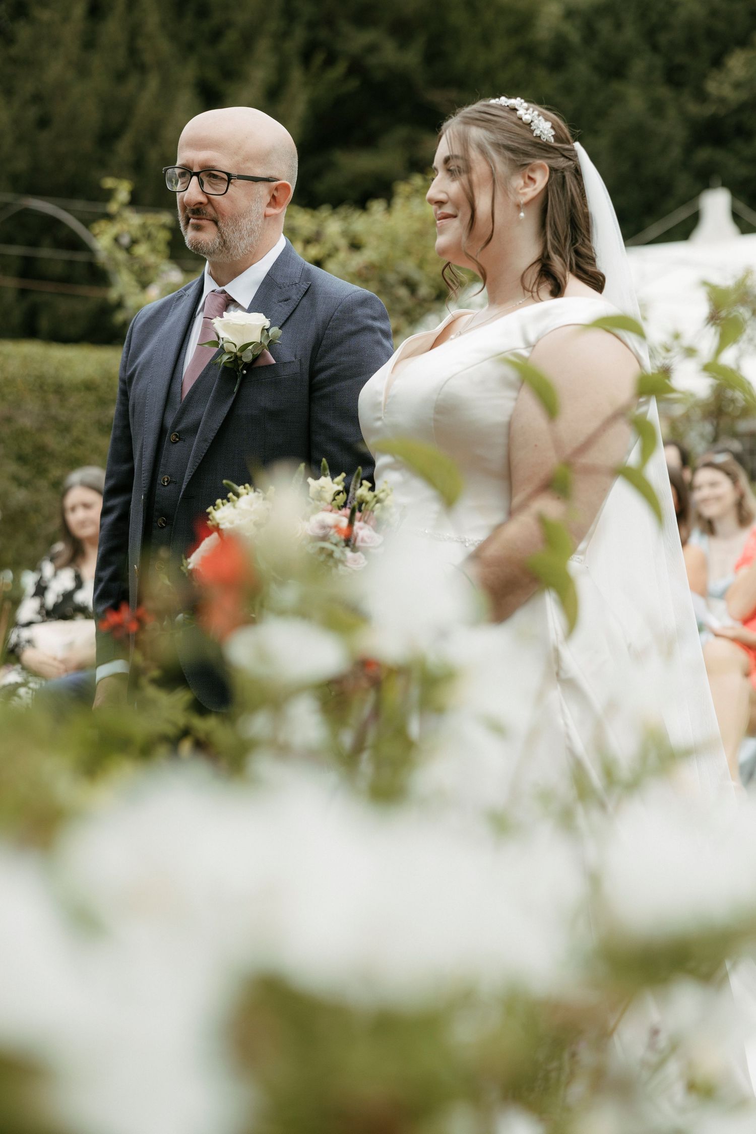 Bride in veil and tiara walking down outdoor aisle with bald bespectacled man in navy suit.