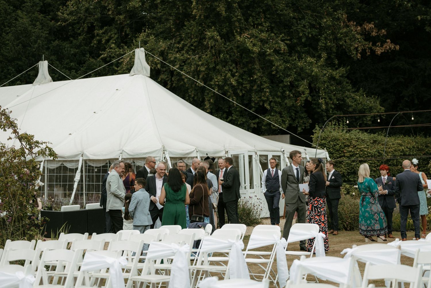 Guests mingle outdoors near a white marquee tent with rows of white chairs set up for a garden wedding ceremony.