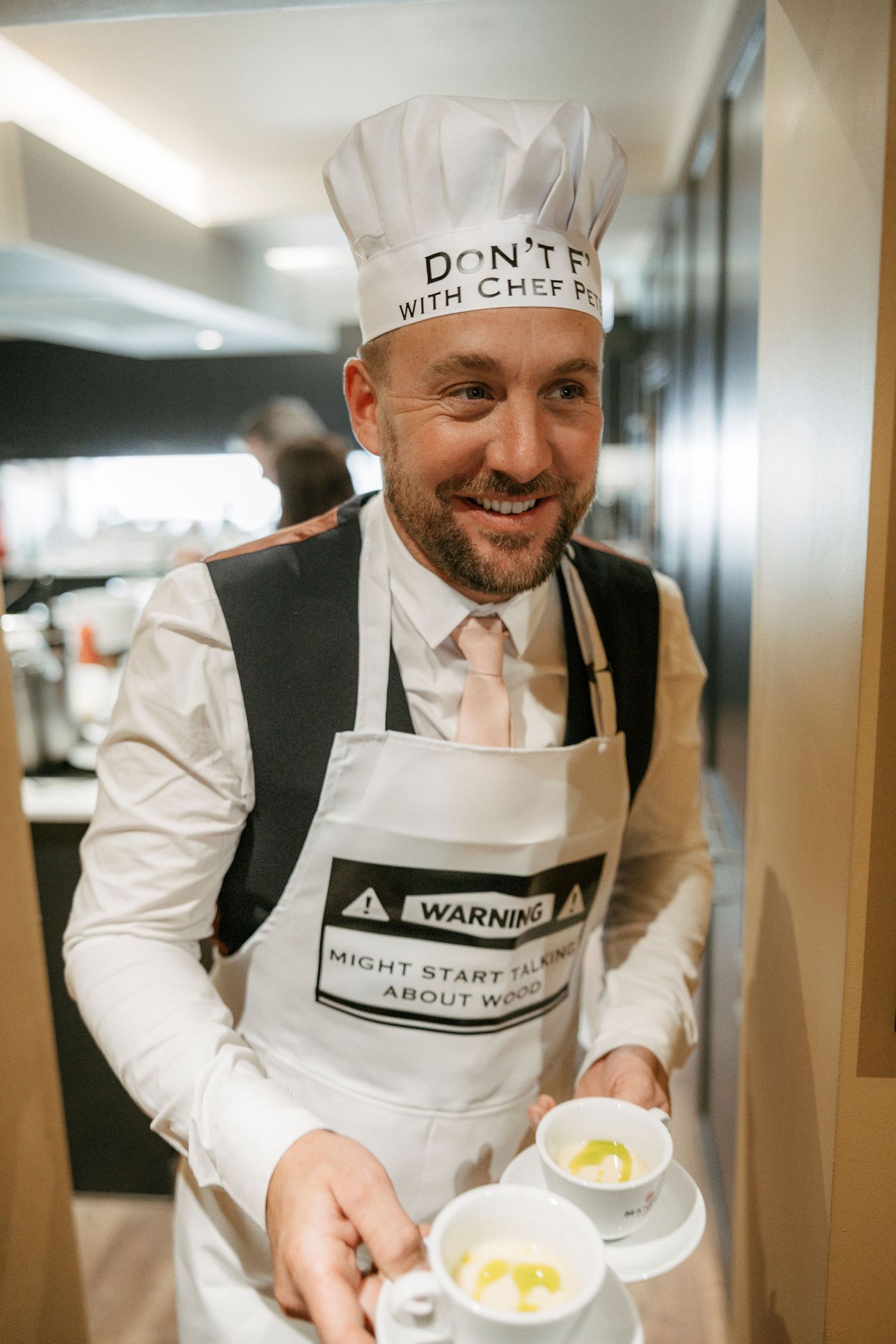 Smiling male chef wearing a humorous chef hat and warning apron holds two small dishes in a modern kitchen.