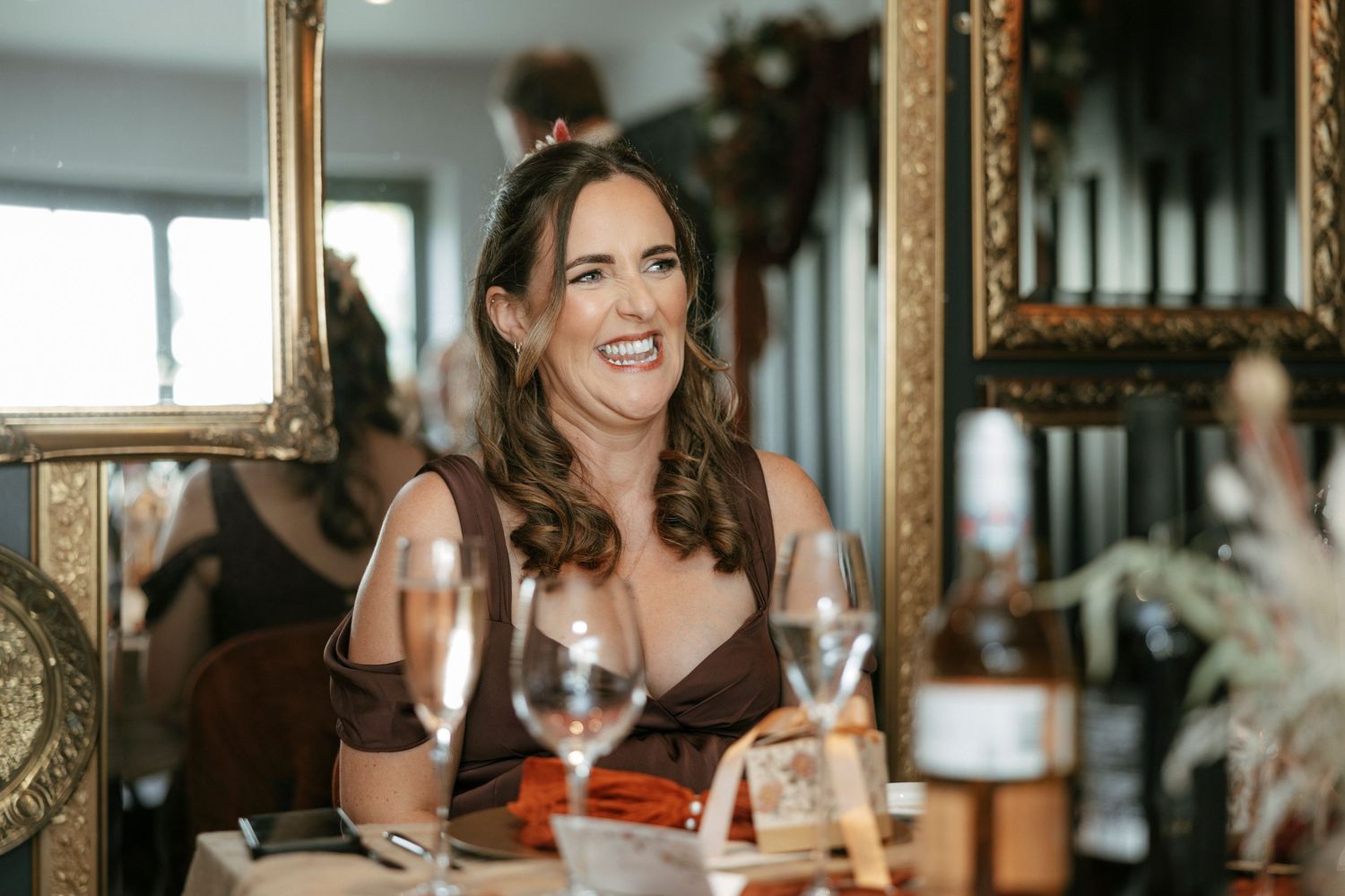 Woman in brown dress laughing at elegant dinner table with wine glasses and gold mirrors.