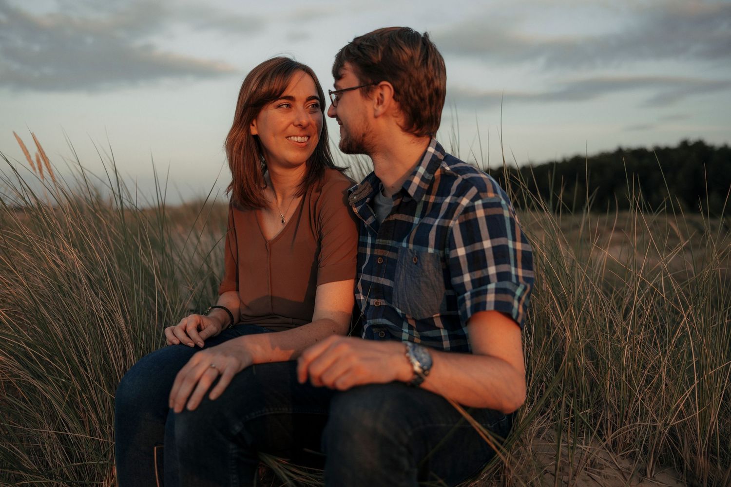 Couple sitting together in grassy dunes at sunset, sharing a romantic moment outdoors.