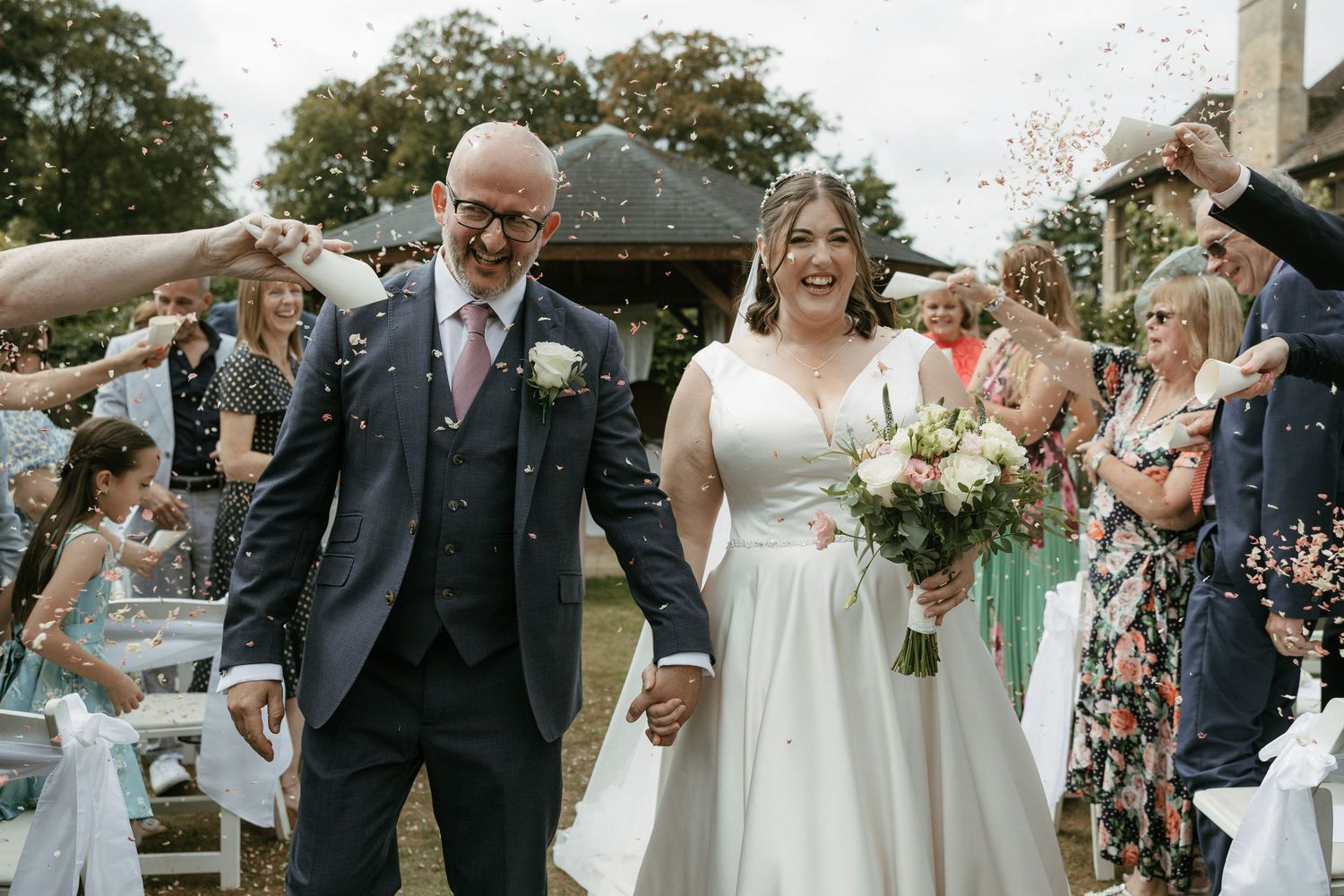 Bride and groom walk hand-in-hand through confetti shower at outdoor wedding ceremony.