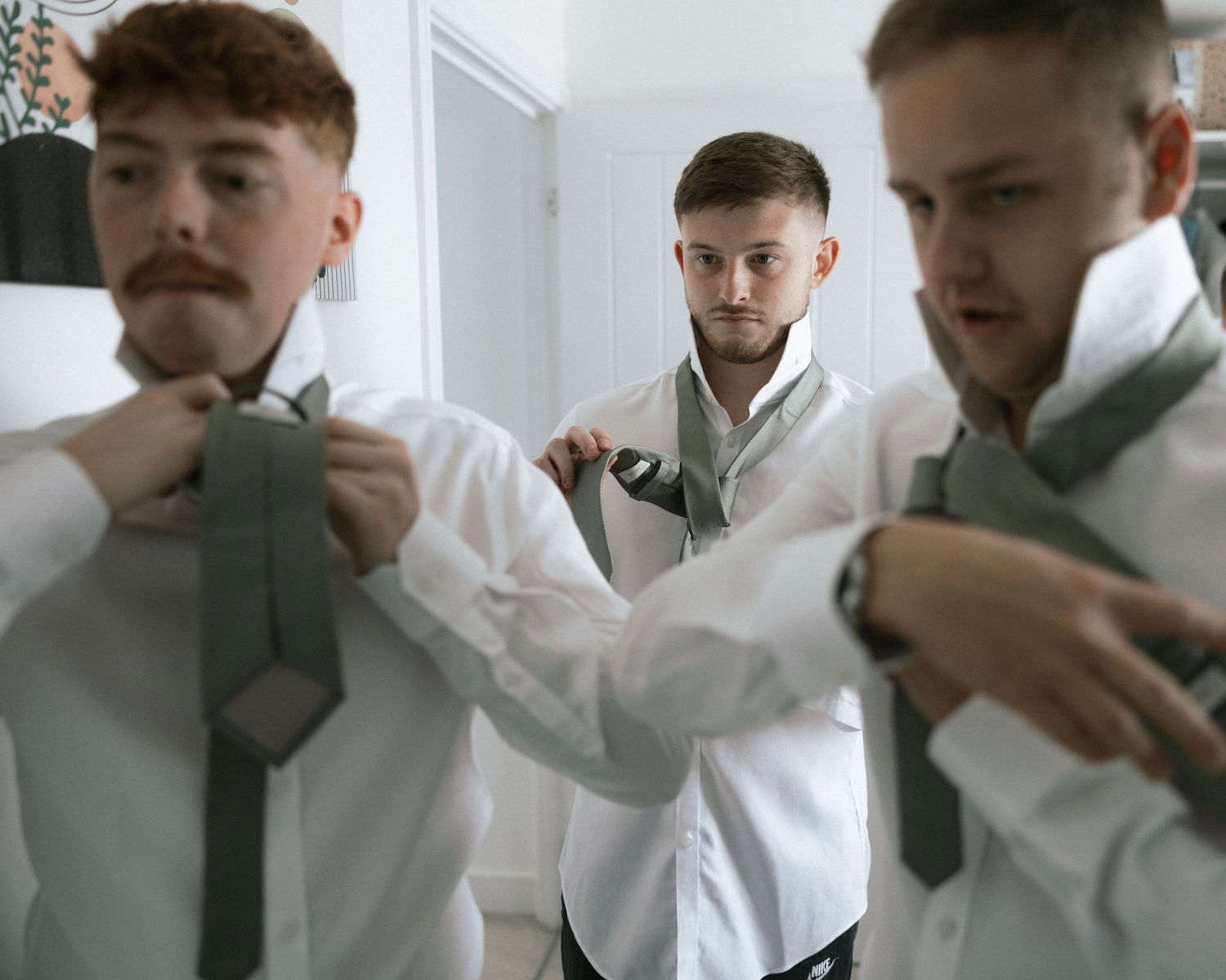Three groomsmen in white shirts adjusting sage green ties before a wedding ceremony.