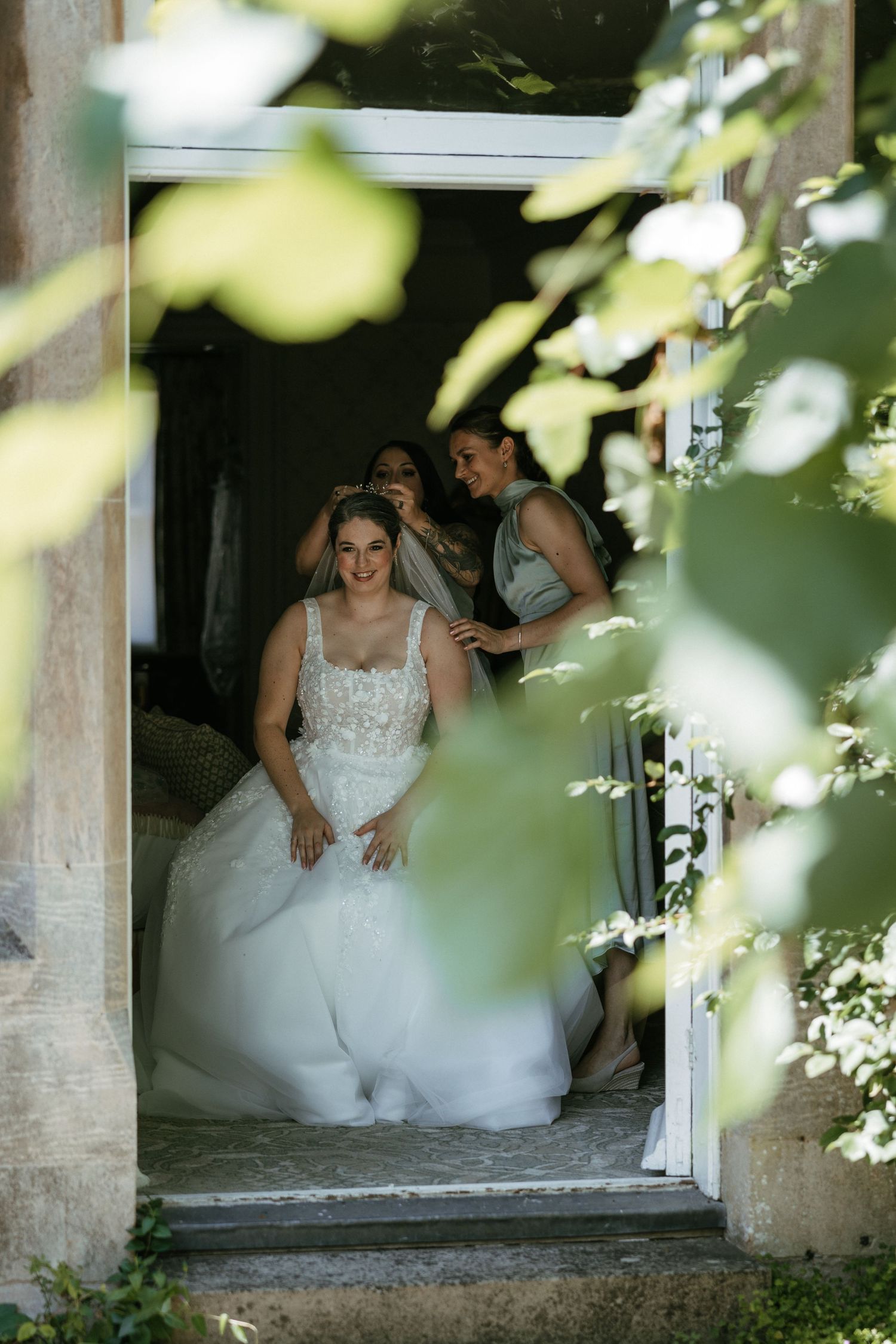 Bride in white gown sits in doorway as bridesmaids adjust her veil, framed by lush green leaves.