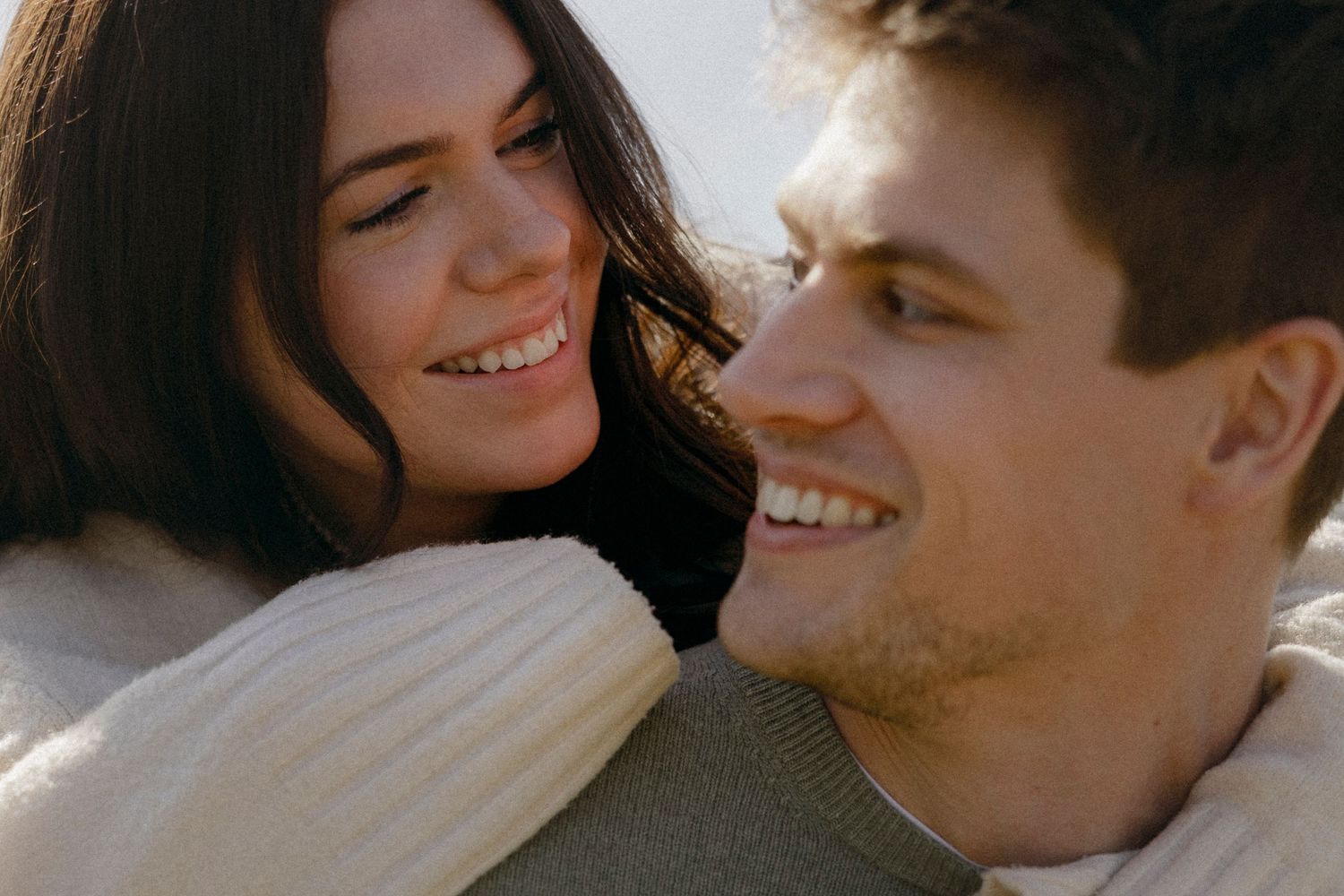 A happy young couple smiling and gazing at each other outdoors, wearing cozy sweaters in soft natural light.