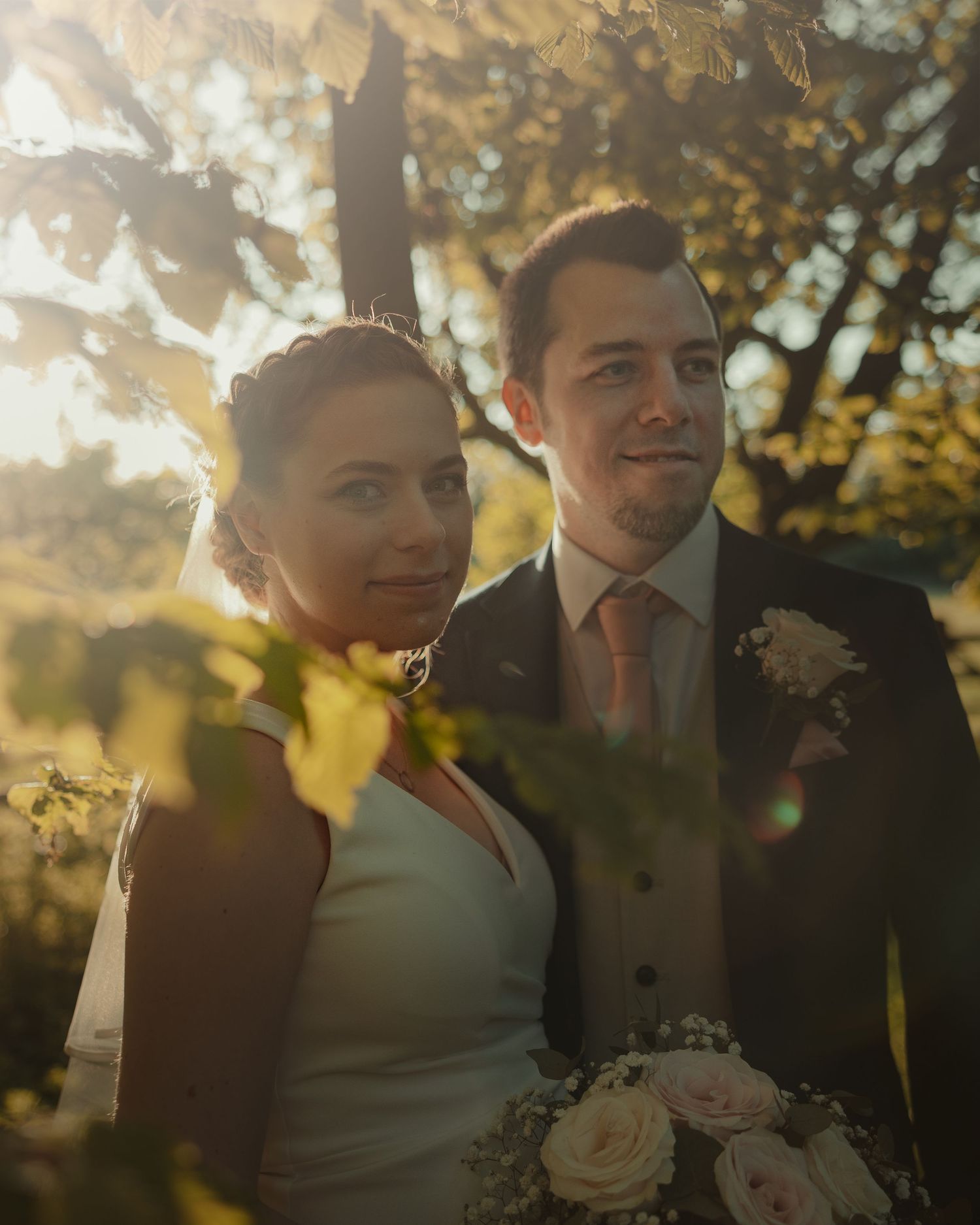 Bride and groom pose together outdoors in golden sunlight, surrounded by autumn foliage on their wedding day.