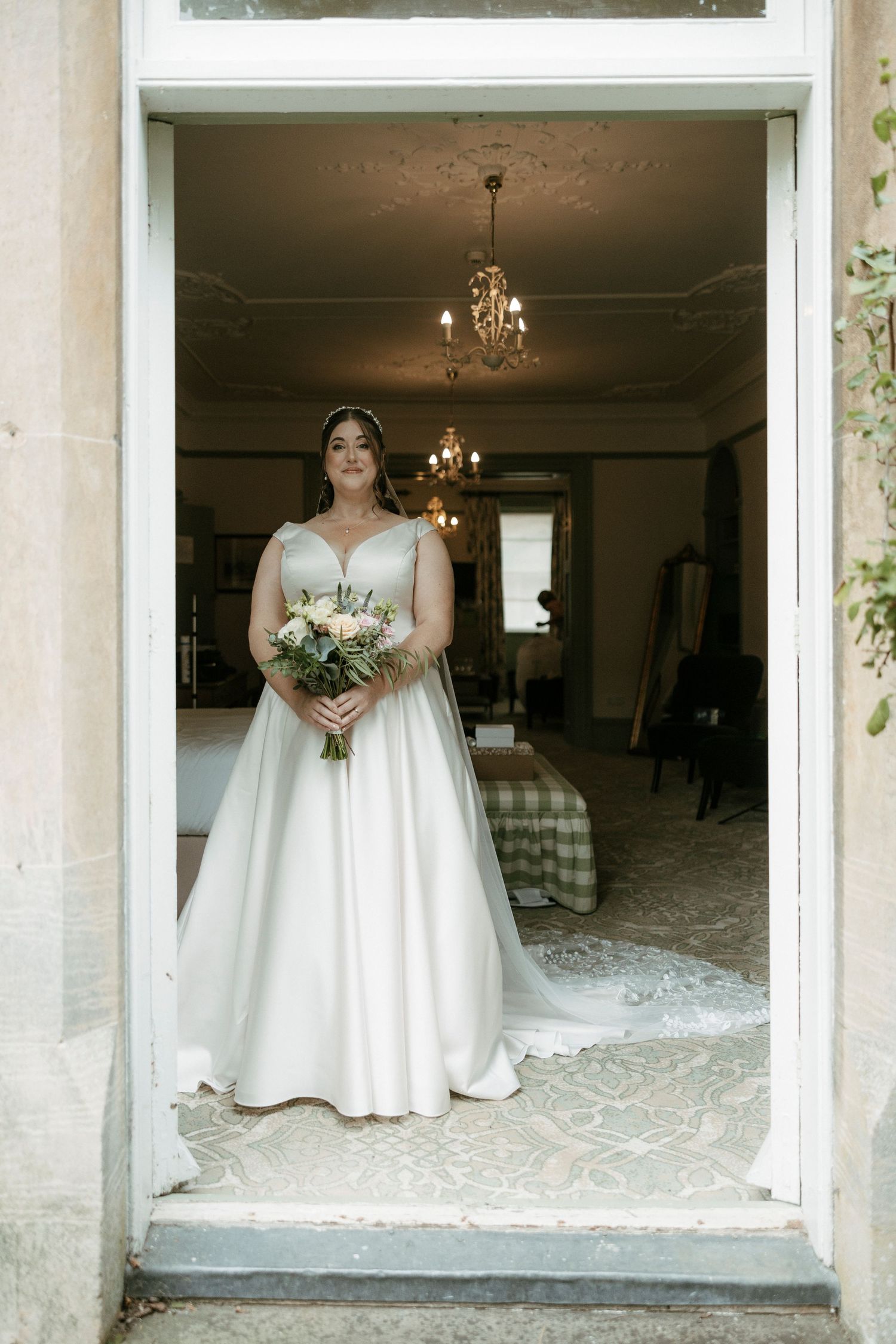 Bride in classic white satin ball gown with V-neck holding floral bouquet standing in doorway of elegant venue.