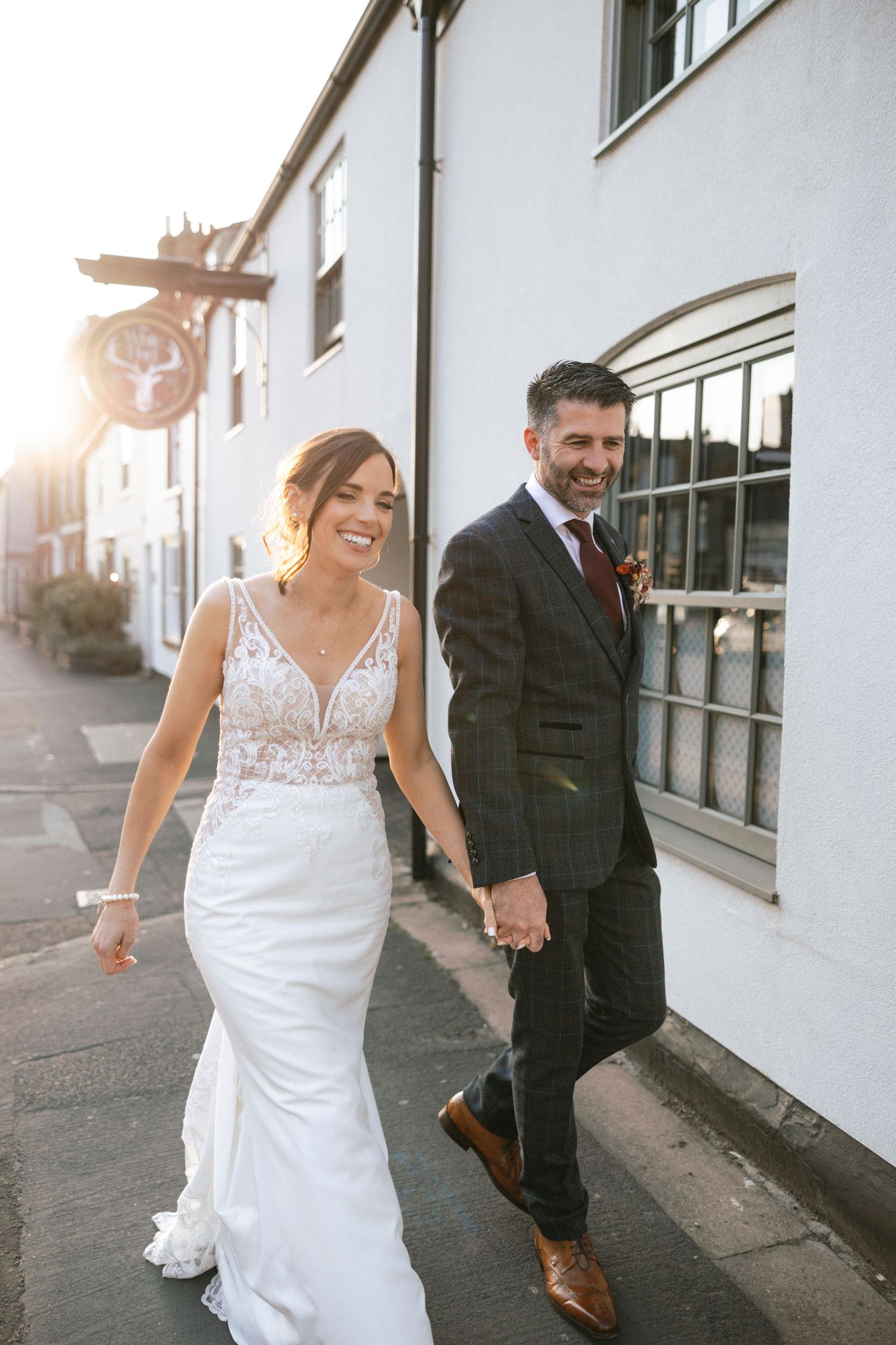 Smiling bride and groom walk hand in hand past a white building in golden sunset light.