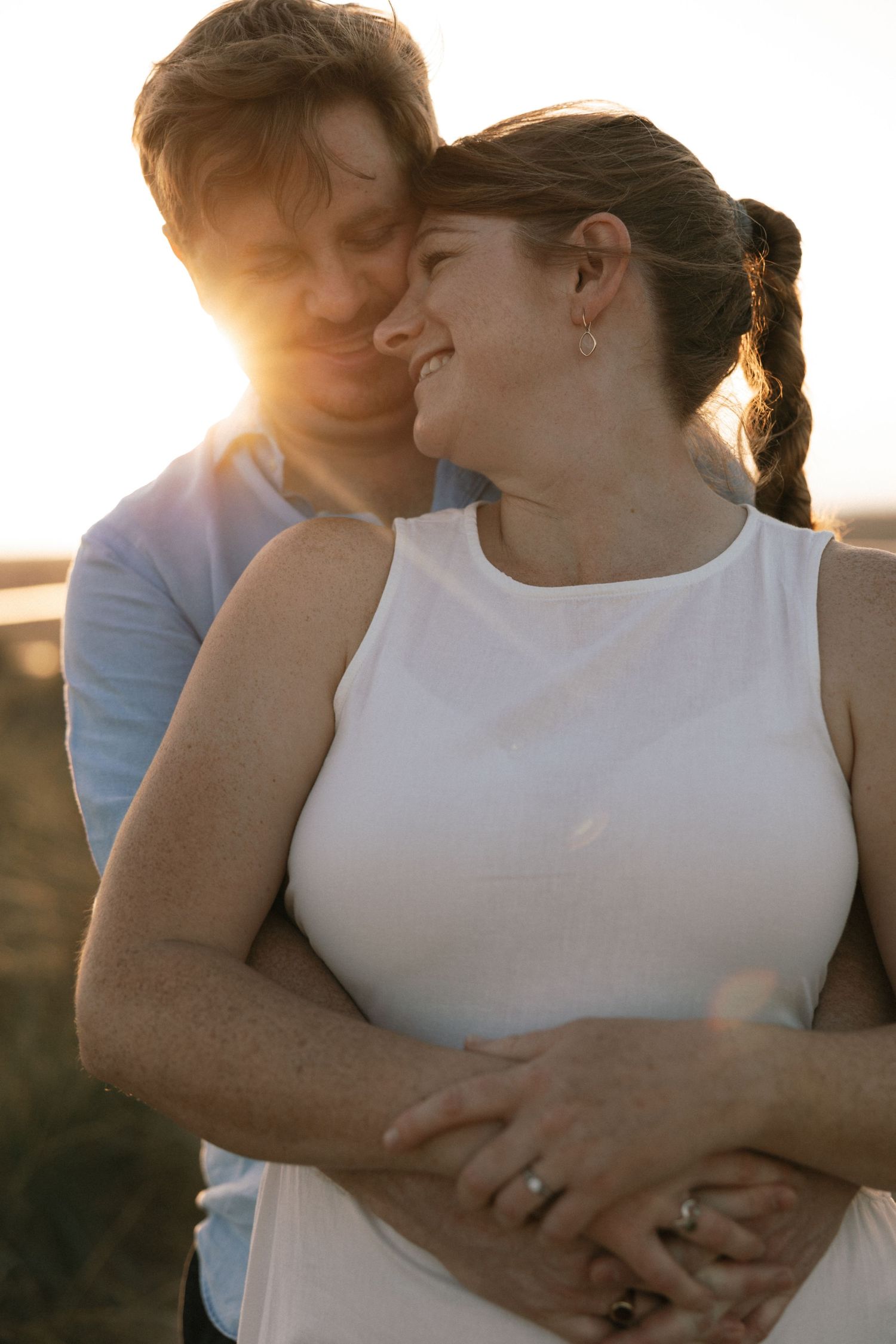A couple embraces at golden hour sunset, woman in white top smiling warmly as man nuzzles her cheek outdoors.