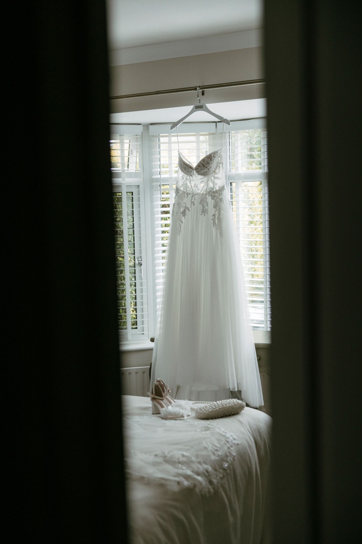 White strapless wedding dress hanging by window with bridal heels on bed below, elegant bridal preparation scene.