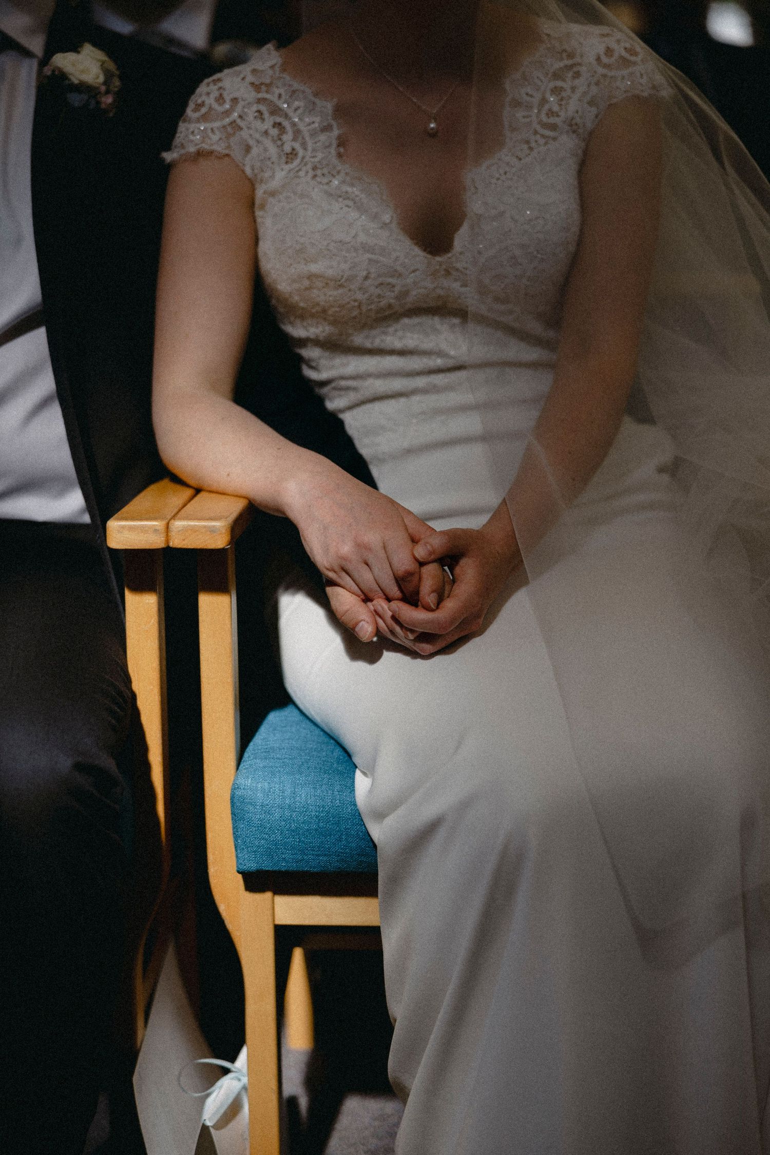 Bride and groom holding hands during wedding ceremony, bride wearing elegant lace white gown while seated on wooden chairs.