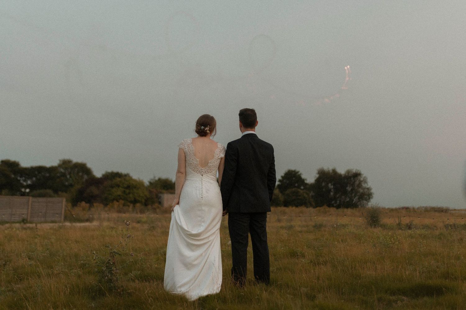 A bride and groom stand together in a field, viewed from behind, wearing wedding attire on a cloudy day.