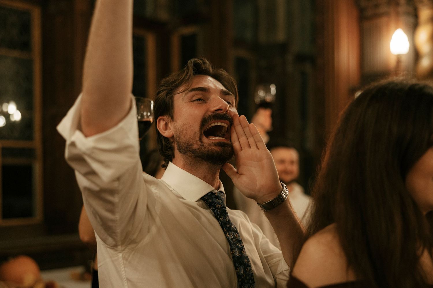 Man in white shirt and tie cheering with arm raised at a lively indoor social gathering or wedding reception.